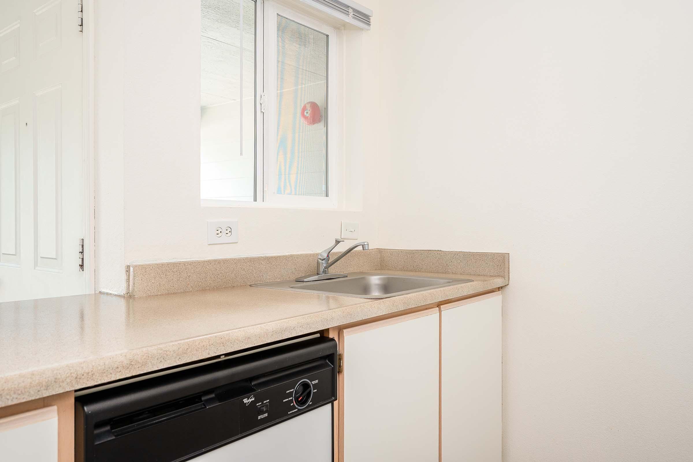 A clean and minimalist kitchen countertop featuring a sink, a dishwasher, and a window with natural light. The walls are painted white, and the cabinetry is a light color, creating a bright and airy atmosphere. The space is uncluttered, emphasizing simplicity.