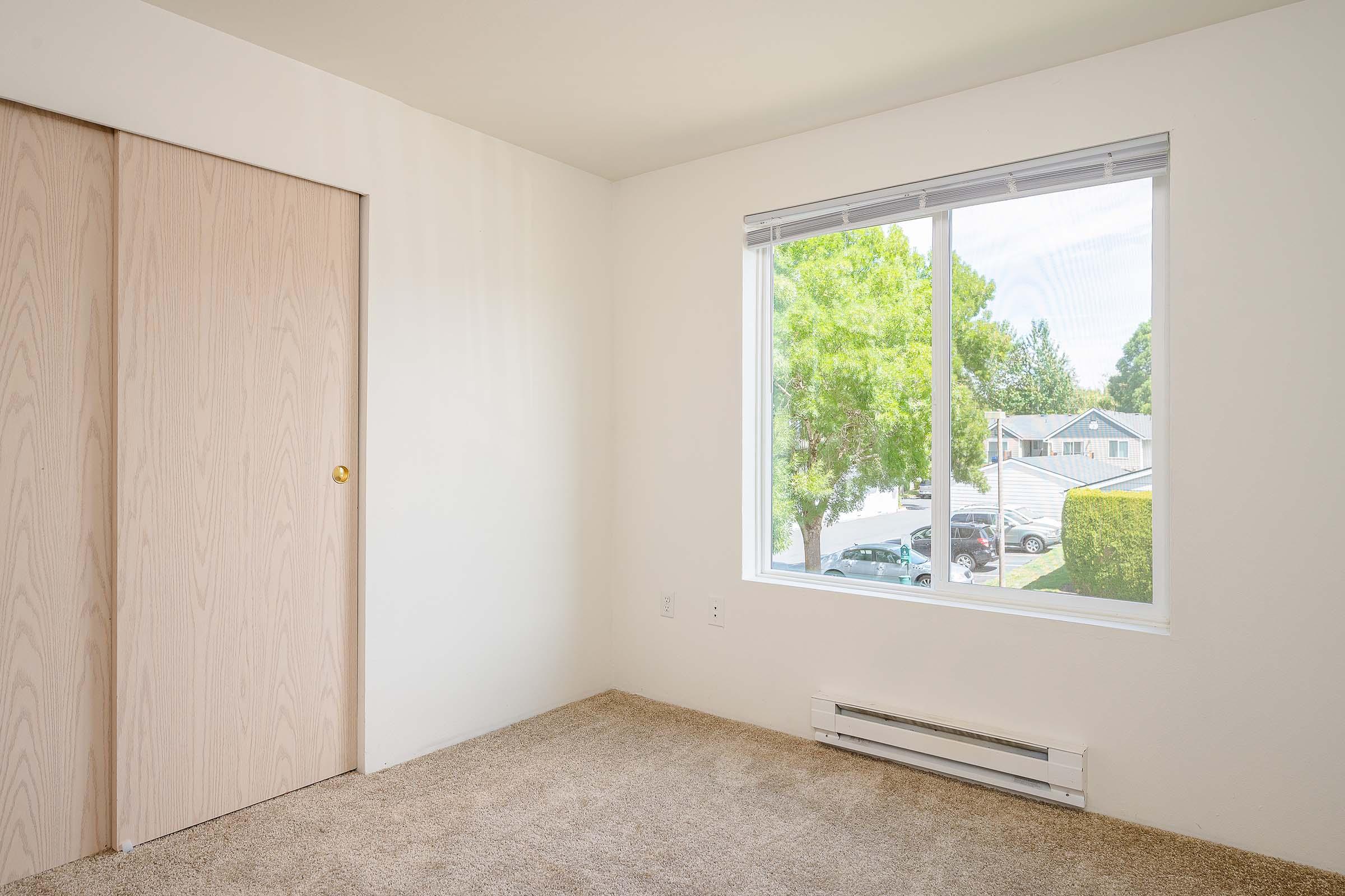 A bright, empty room featuring light-colored walls and a window overlooking a residential area. The floor is carpeted, and there is a wooden closet door on one side. Sunlight filters in, creating a warm and inviting atmosphere.