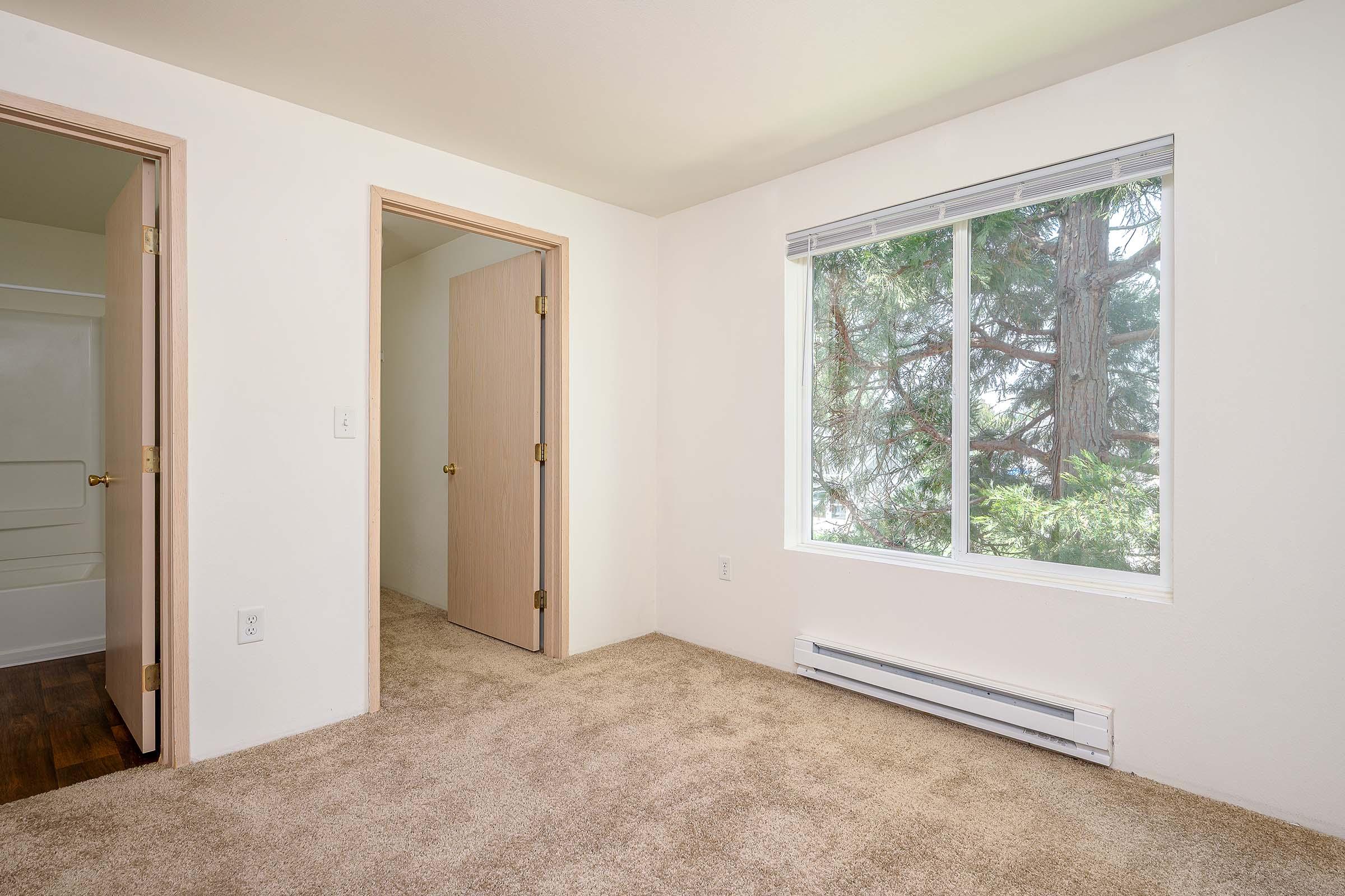A spacious empty room with light beige carpet, featuring two doors; one leading to a bathroom and the other to a hallway. A large window allows natural light to fill the space, providing a view of greenery outside. The walls are painted in a light color, enhancing the bright and airy feel of the room.