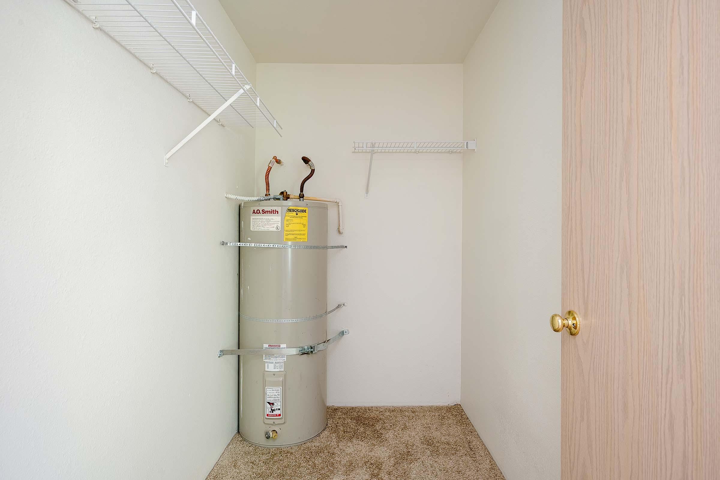 A small storage closet featuring a water heater along one wall, shelves mounted on adjacent walls, and a closed wooden door. The room has light-colored walls and a carpeted floor.