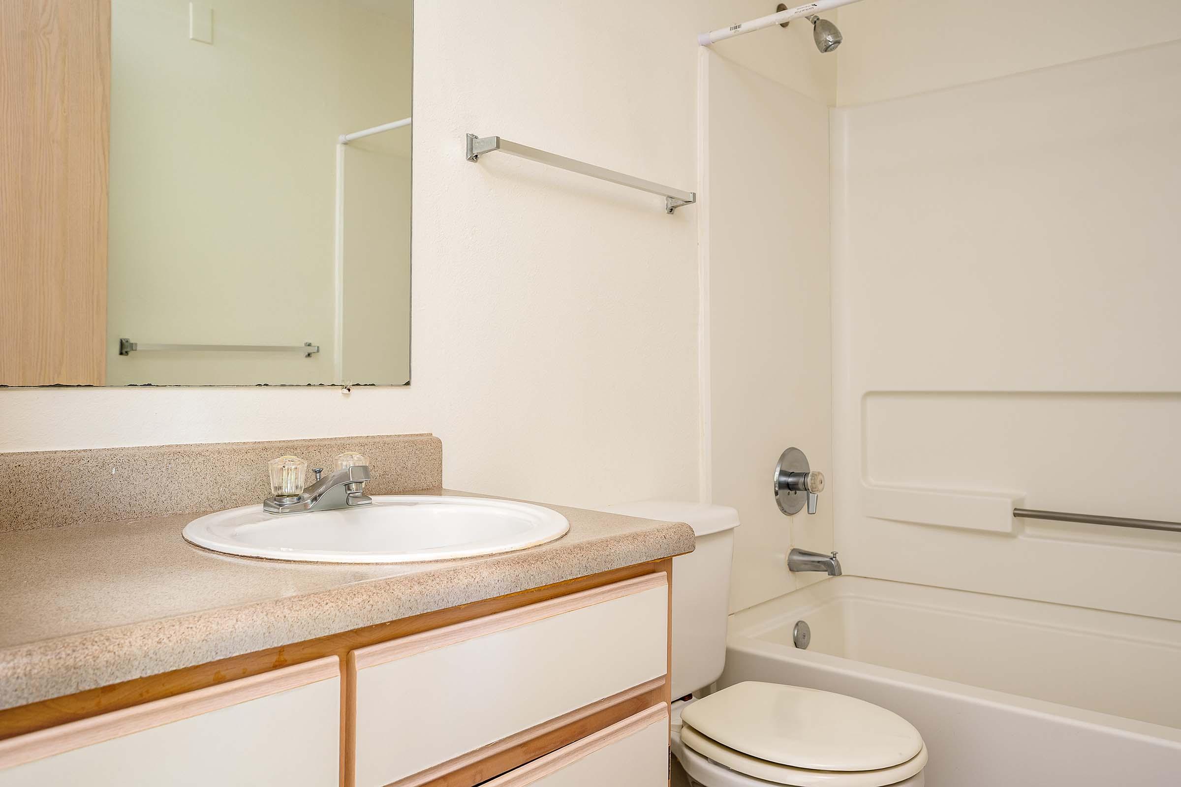 A clean, minimalistic bathroom featuring a white bathtub with a shower, a sink with a round basin, and a small mirror above it. The walls are painted in a light color, and there are wooden cabinets beneath the sink, providing storage space. Natural light fills the room.