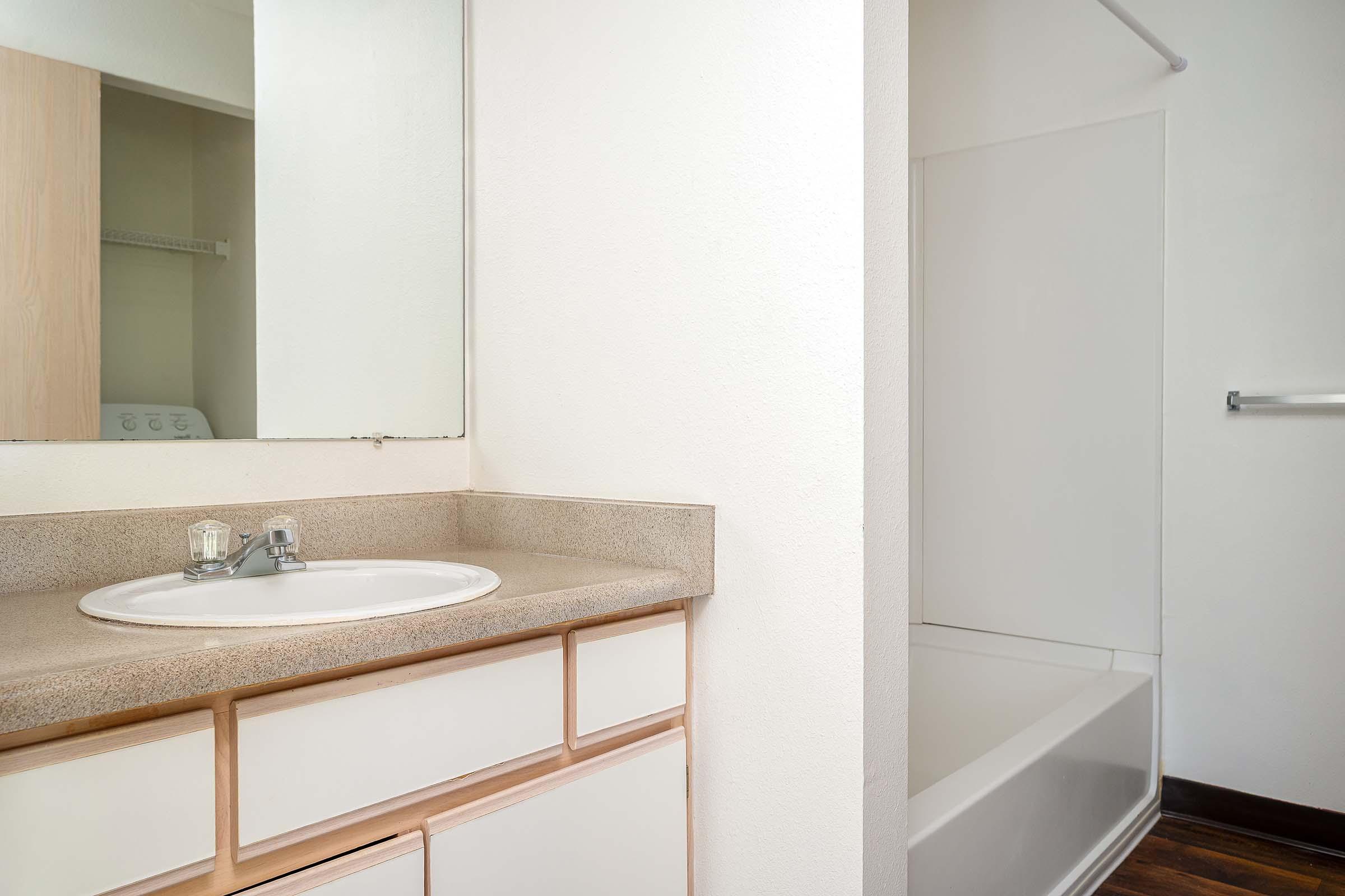 Bathroom interior featuring a sink with a mirror above, a countertop, and a bathtub with a showerhead. The walls are painted white, and the floor appears to be wooden. A laundry area is visible in the background.
