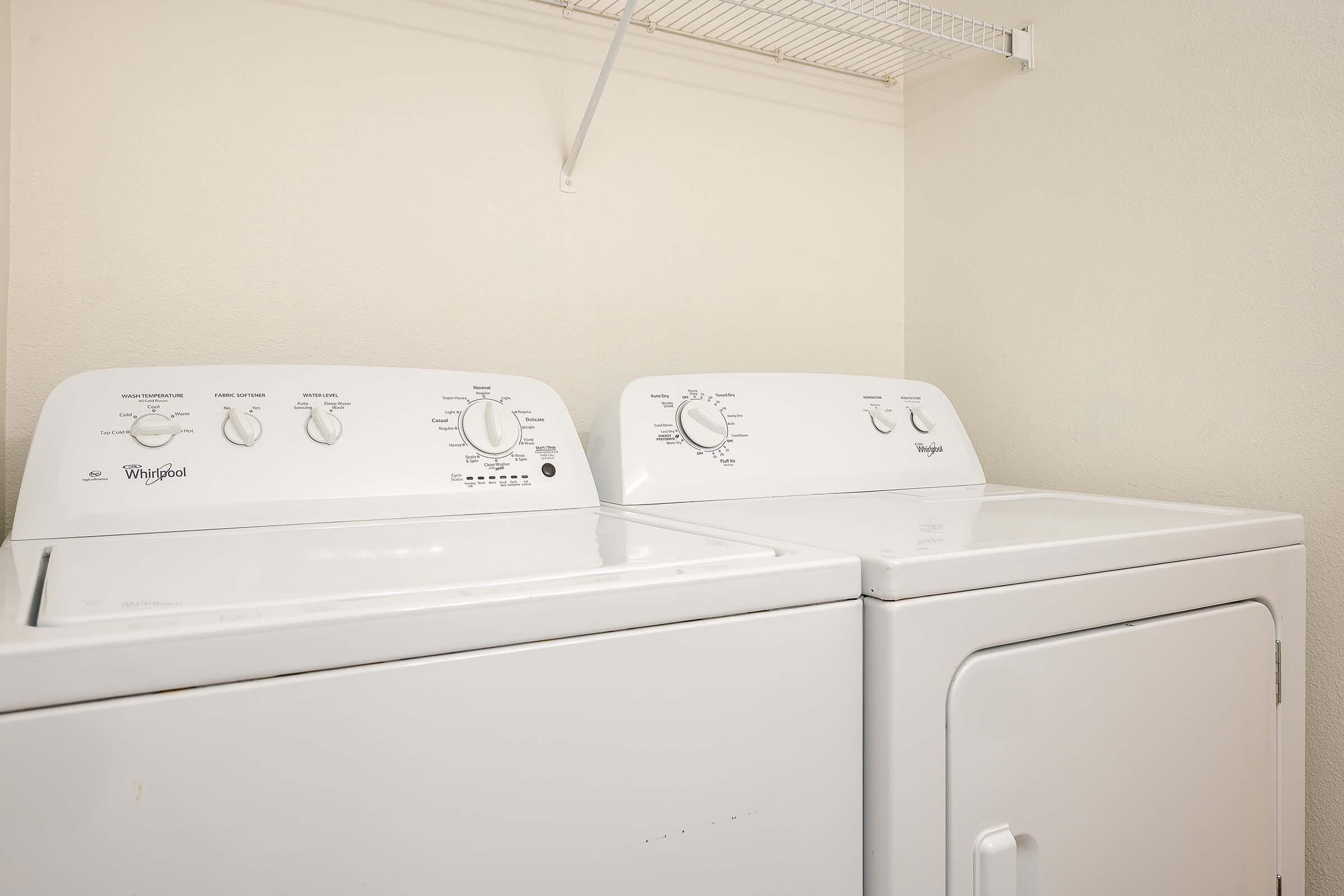 A white washing machine and dryer side by side in a laundry area, with control dials and settings visible. The machines are positioned against a plain wall, and there's a metal shelf above them for storage. The overall setting is bright and clean.