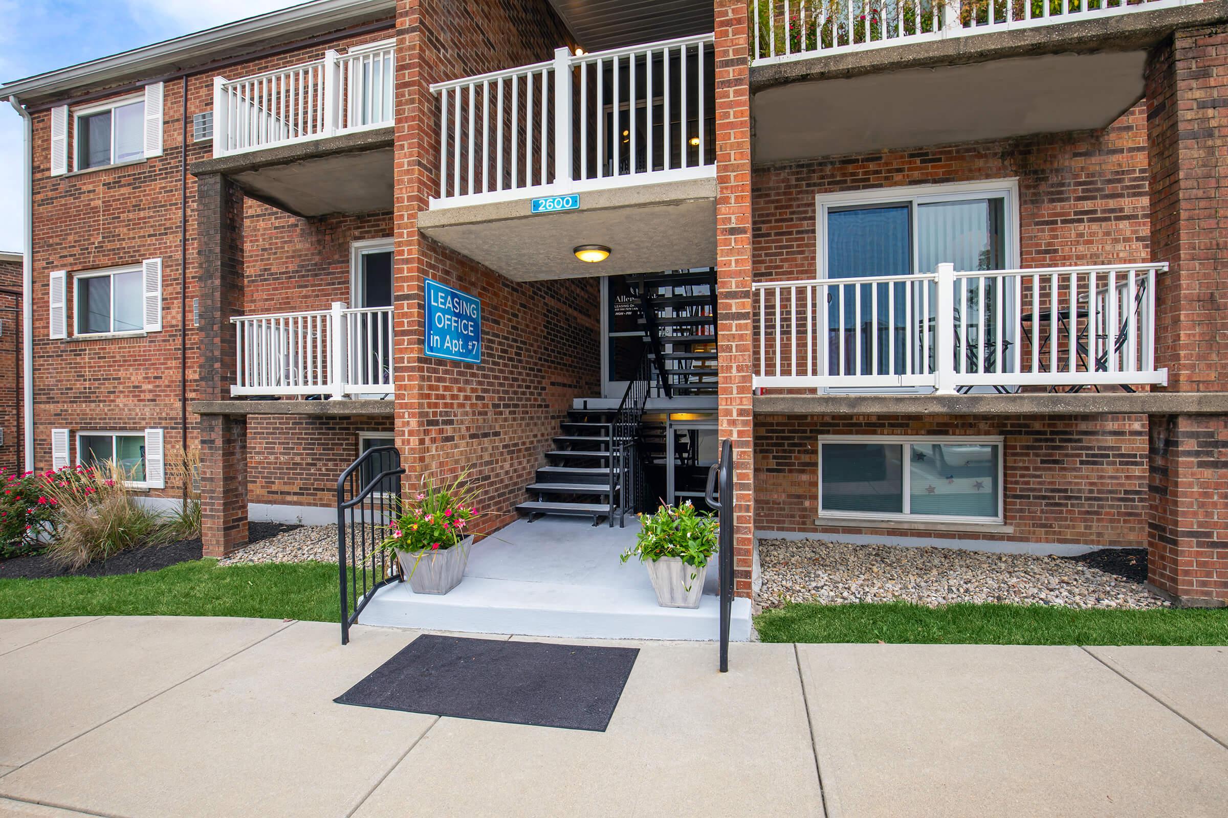 Exterior view of an apartment building featuring a leasing office entrance. The entrance has a set of stairs leading up to the second floor, flanked by flower planters. A blue sign indicates the leasing office, and there are balconies on the upper level. The landscaping includes neatly maintained grass and decorative stones.
