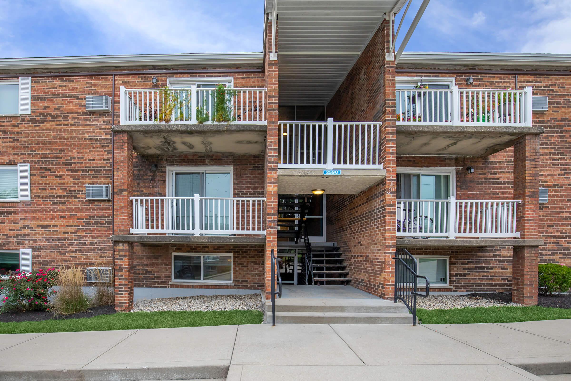 A two-story brick apartment building featuring balconies on both levels. The entrance has a central staircase leading to upper units. Lush landscaping with flowers borders the walkway and ground level, with clear blue skies overhead.