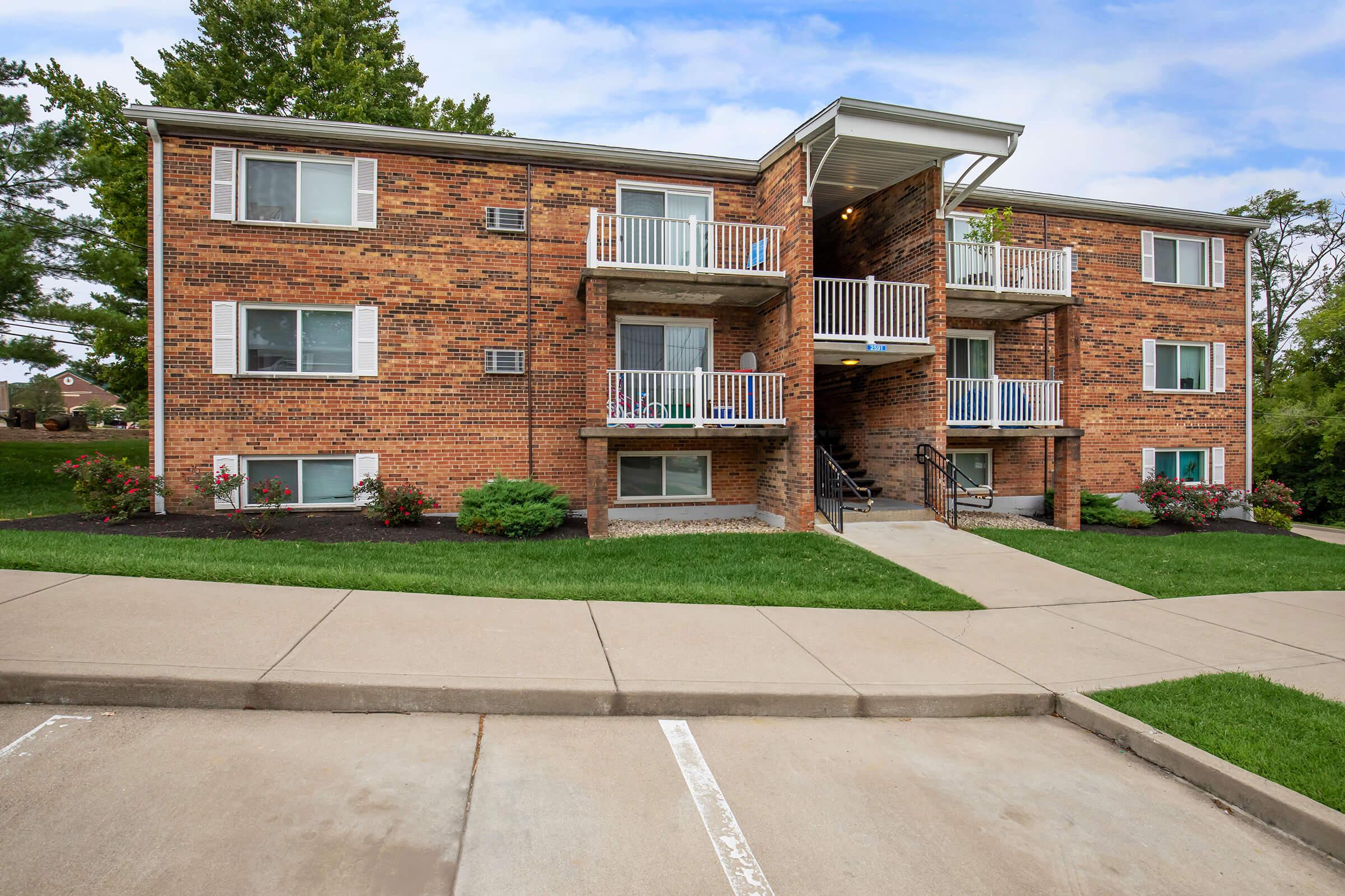 A brick apartment building with three stories, featuring multiple balconies on each level. Well-maintained landscaping surrounds the building, with green grass and flower beds. A concrete pathway leads to the entrance, and a parking area is visible in the foreground.