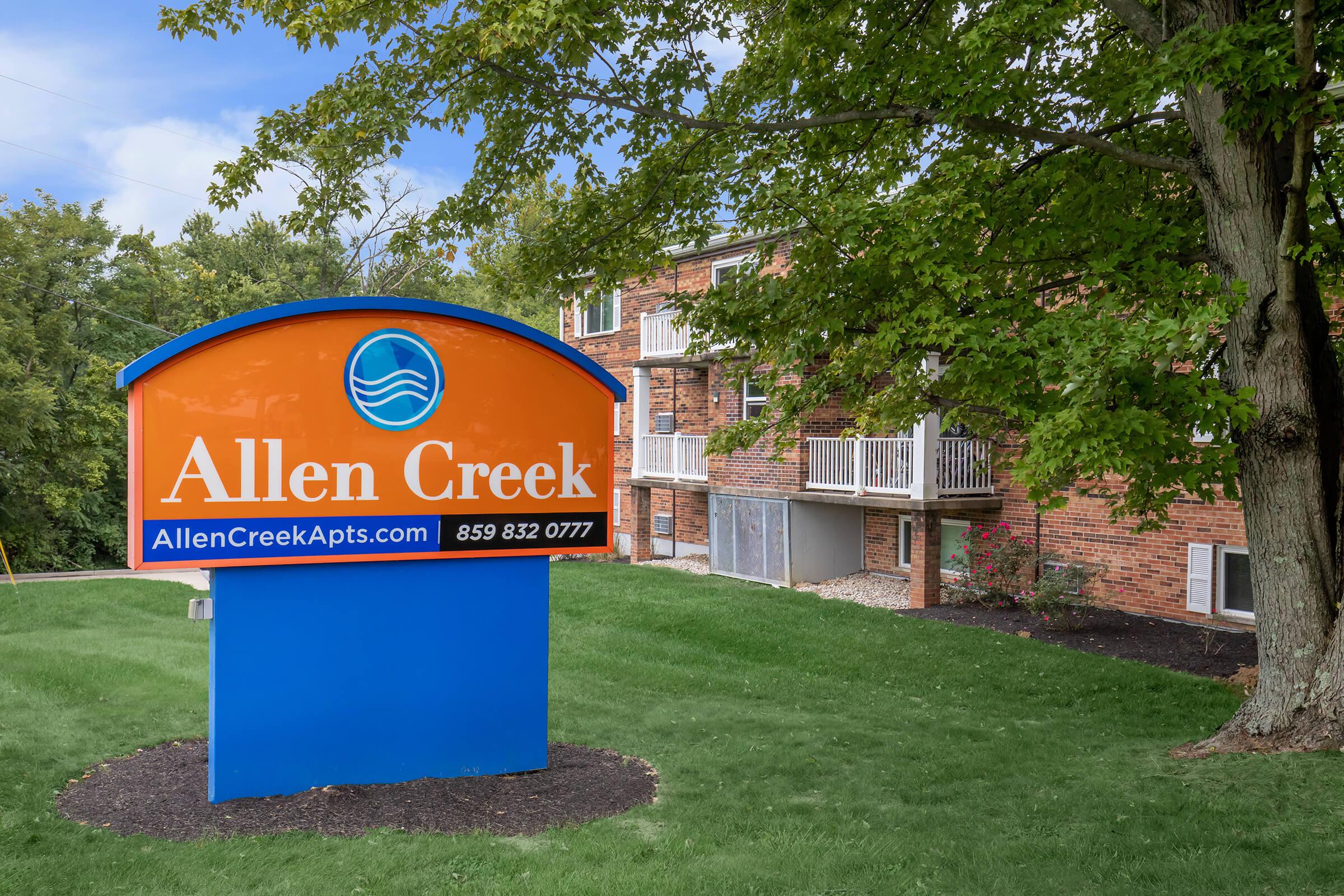 Sign for Allen Creek Apartments featuring an orange and blue color scheme. The sign includes the name "Allen Creek," a website URL (AllenCreekApts.com), and a phone number (859-832-0777). Surrounding greenery and a brick building are visible in the background.