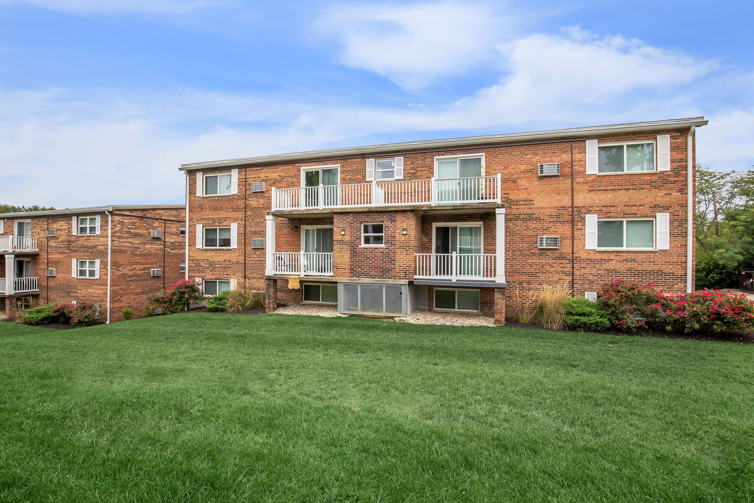 A brick apartment building with multiple units, featuring balconies and air conditioning units. The building is surrounded by a green lawn and colorful flower bushes, under a clear blue sky.