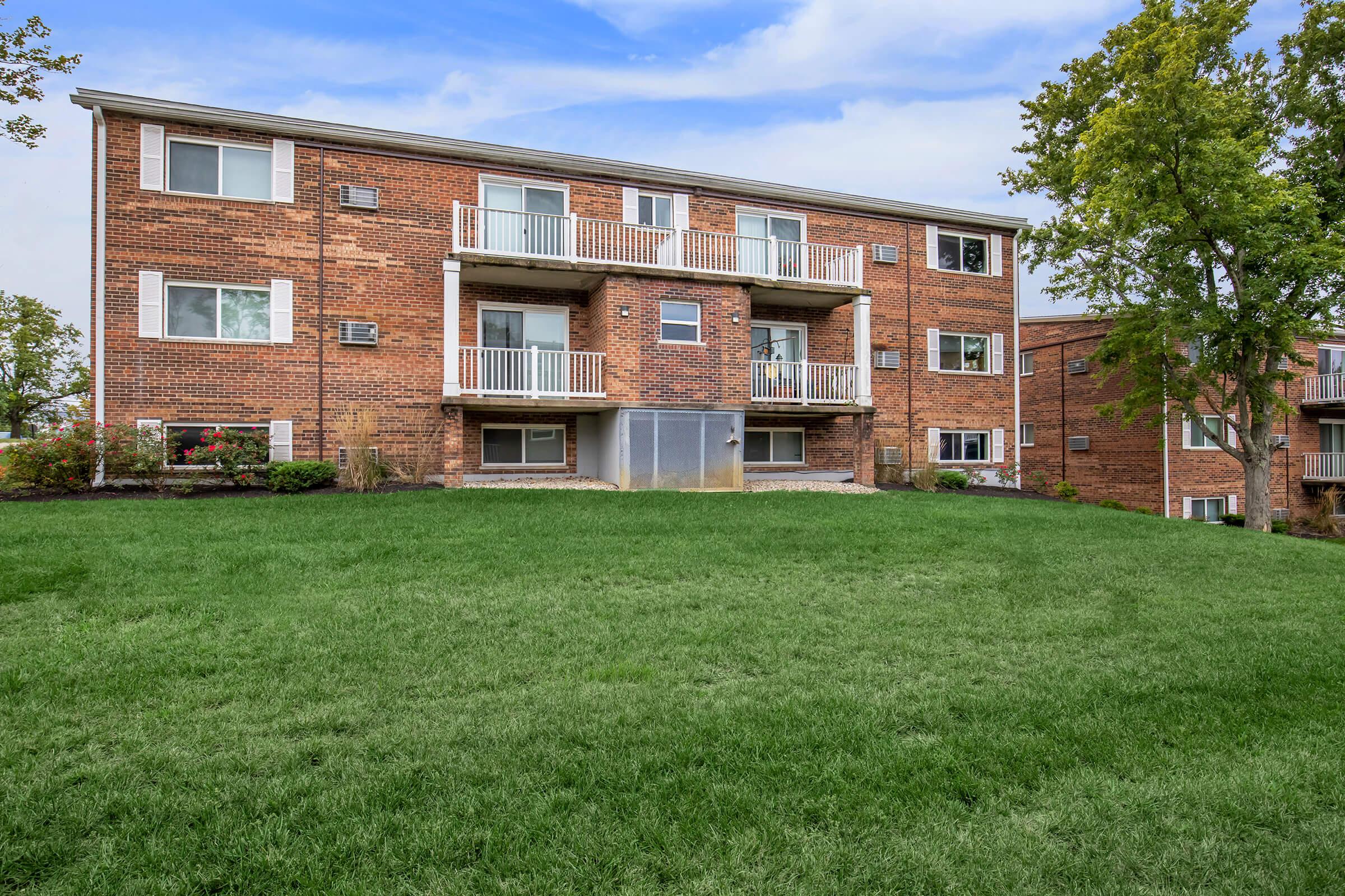 A multi-unit brick apartment building with balconies on the second floor, set against a grassy lawn and surrounded by trees. The sky is partly cloudy, and the scene conveys a residential environment.