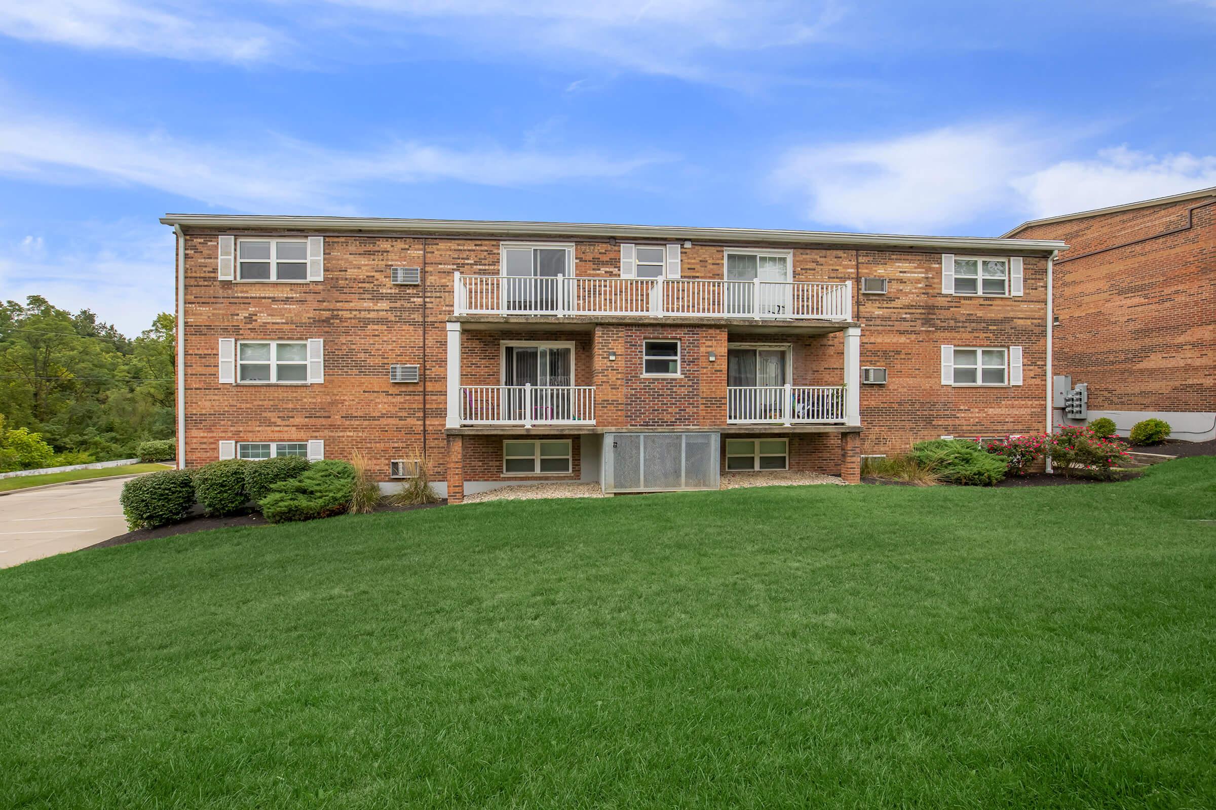 A multistory brick apartment building with balconies, situated on a grassy hillside. The building features several windows and landscaped shrubs in the foreground, under a clear blue sky.
