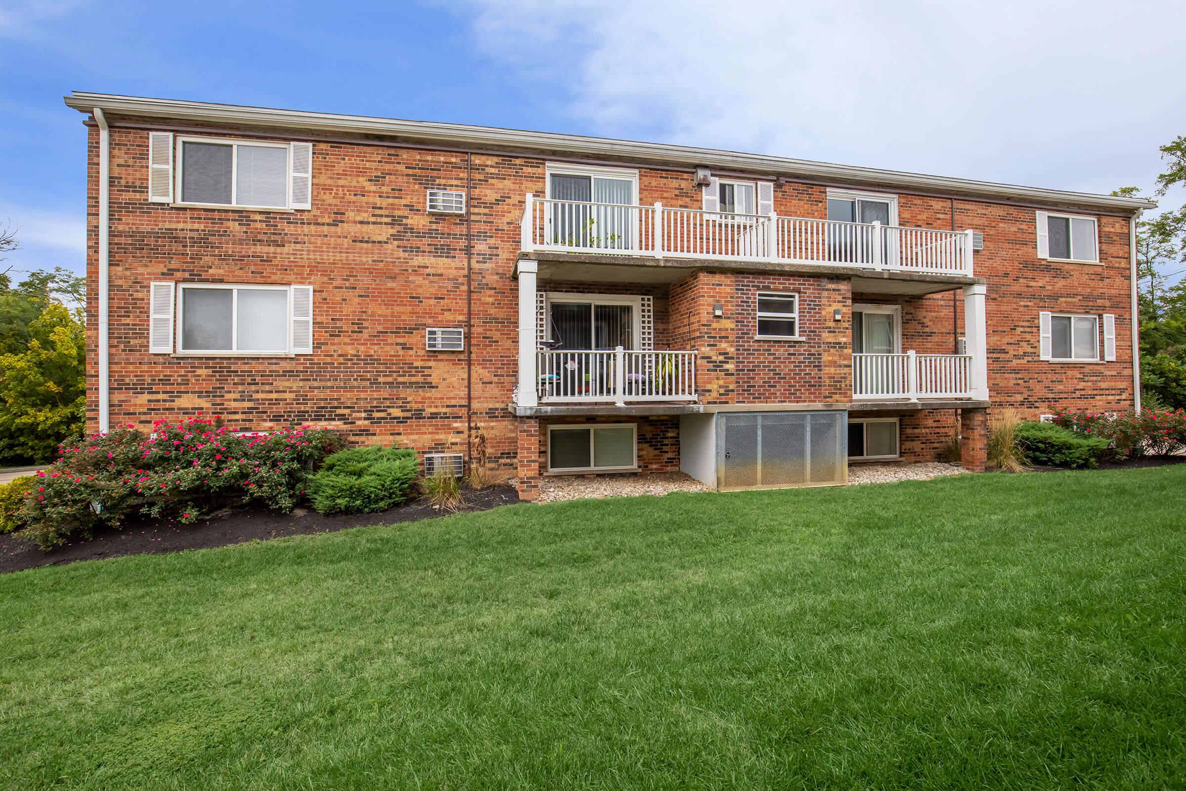 A two-story brick apartment building featuring multiple windows and two balconies on the second floor. The building is surrounded by well-maintained grass and flower beds, with a clear blue sky above.