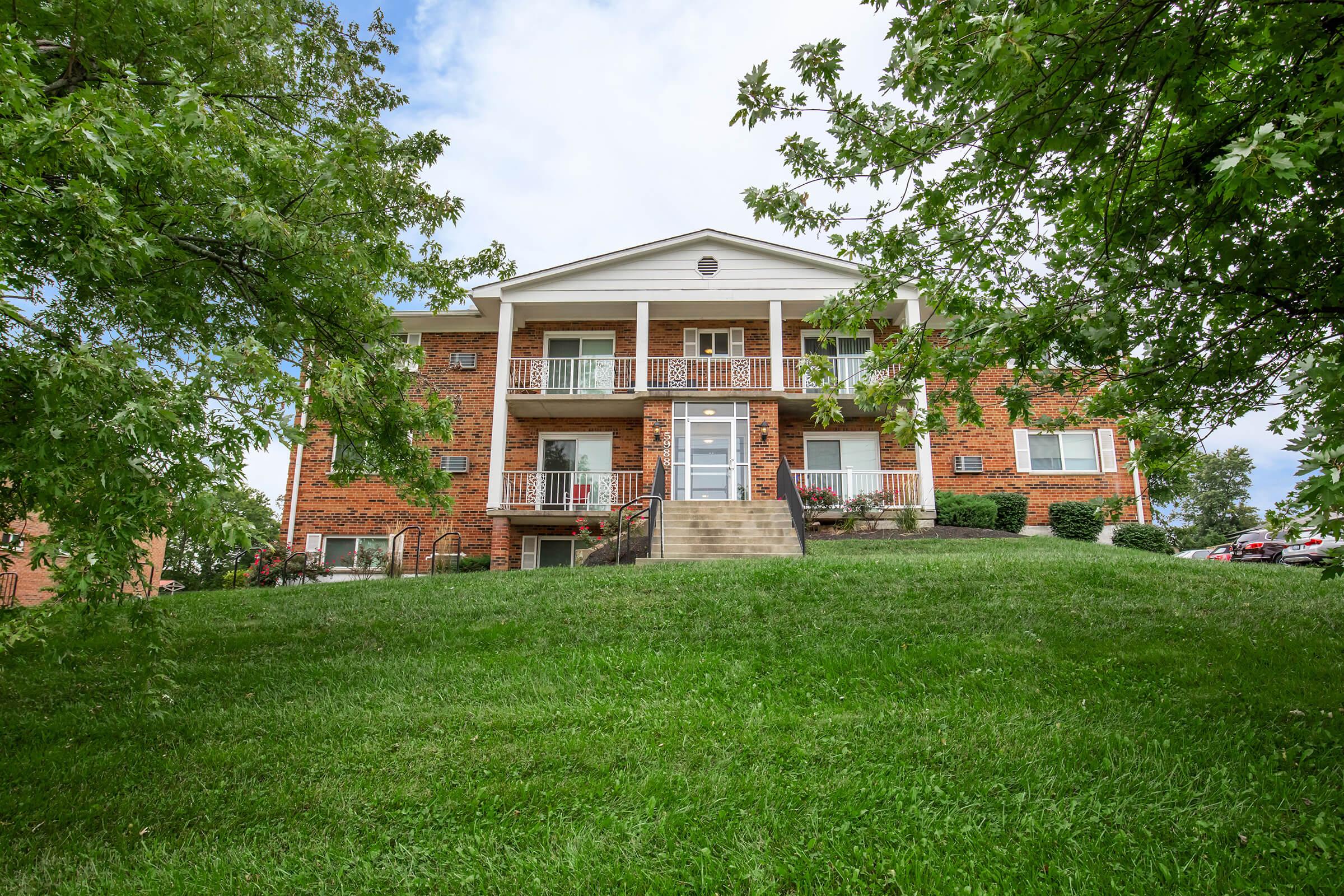 A three-story brick apartment building set on a hillside, surrounded by green grass and trees. The building features white balconies and a staircase leading to the entrance. The sky is partly cloudy, creating a pleasant suburban atmosphere.