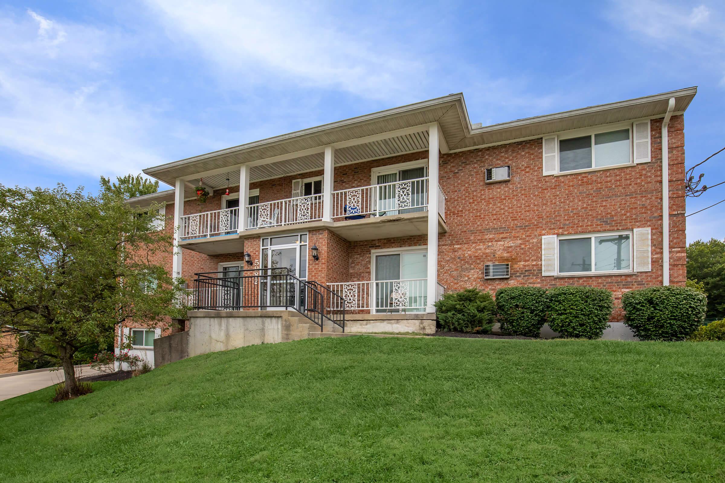 A two-story brick apartment building with a front staircase leading to the main entrance. There are balconies on both levels, and the property features a green lawn in front. Decorative plants are visible on the balconies, and the sky is clear with a few clouds.