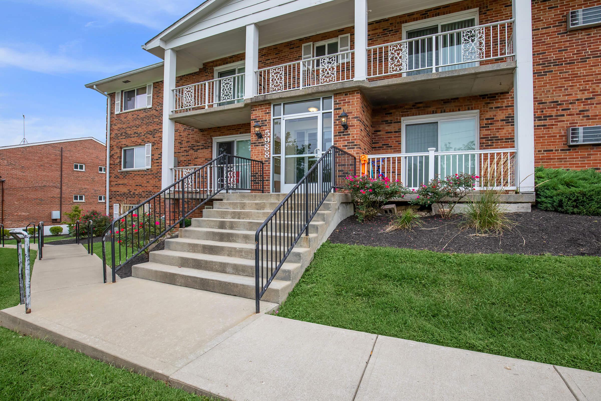 A brick apartment building with a set of stairs leading up to the main entrance. The entrance features glass doors and is flanked by flower beds. The surrounding area has well-maintained grass and sidewalks, with additional buildings visible in the background under a blue sky.