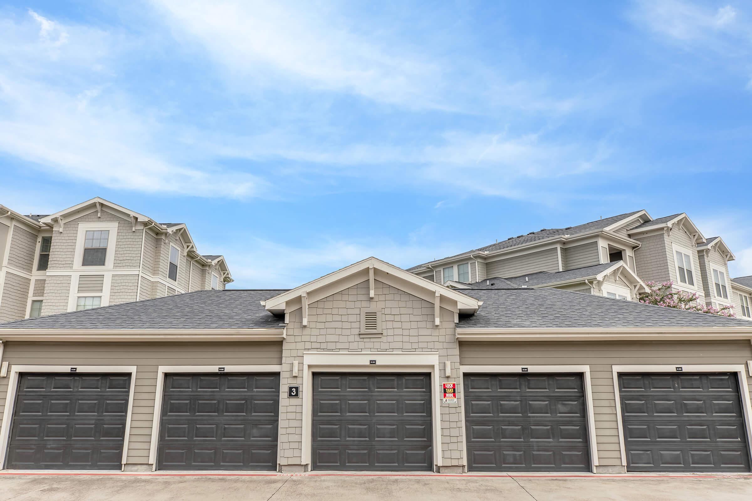 A row of three garage doors in a residential area, with a light gray exterior building in the background and a clear blue sky above. The garages have dark gray doors, and the scene conveys a calm suburban atmosphere.