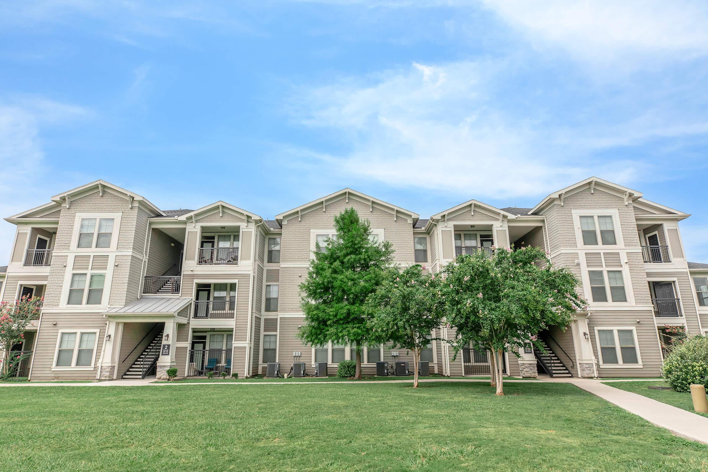 A three-story residential apartment building with a beige exterior, featuring multiple balconies and staircases. In the foreground, there are two small trees and a well-maintained green lawn. The sky is clear and blue, creating a pleasant atmosphere.