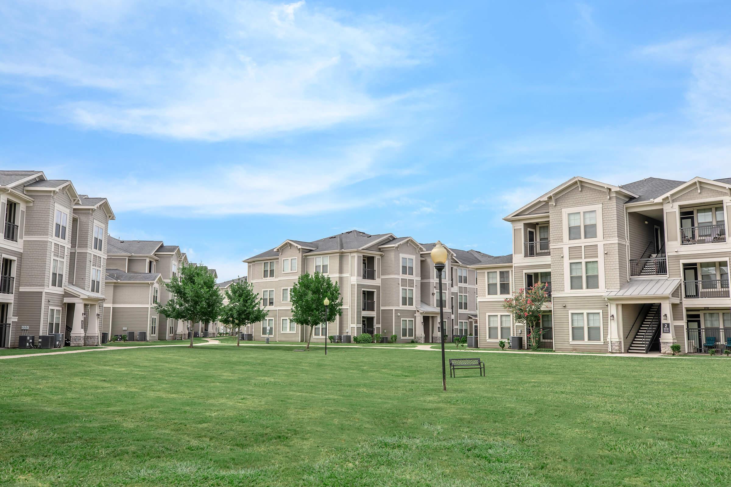 A spacious green courtyard surrounded by modern apartment buildings under a blue sky. The area features well-maintained lawns and trees, with benches and pathways visible, creating a welcoming outdoor living space.