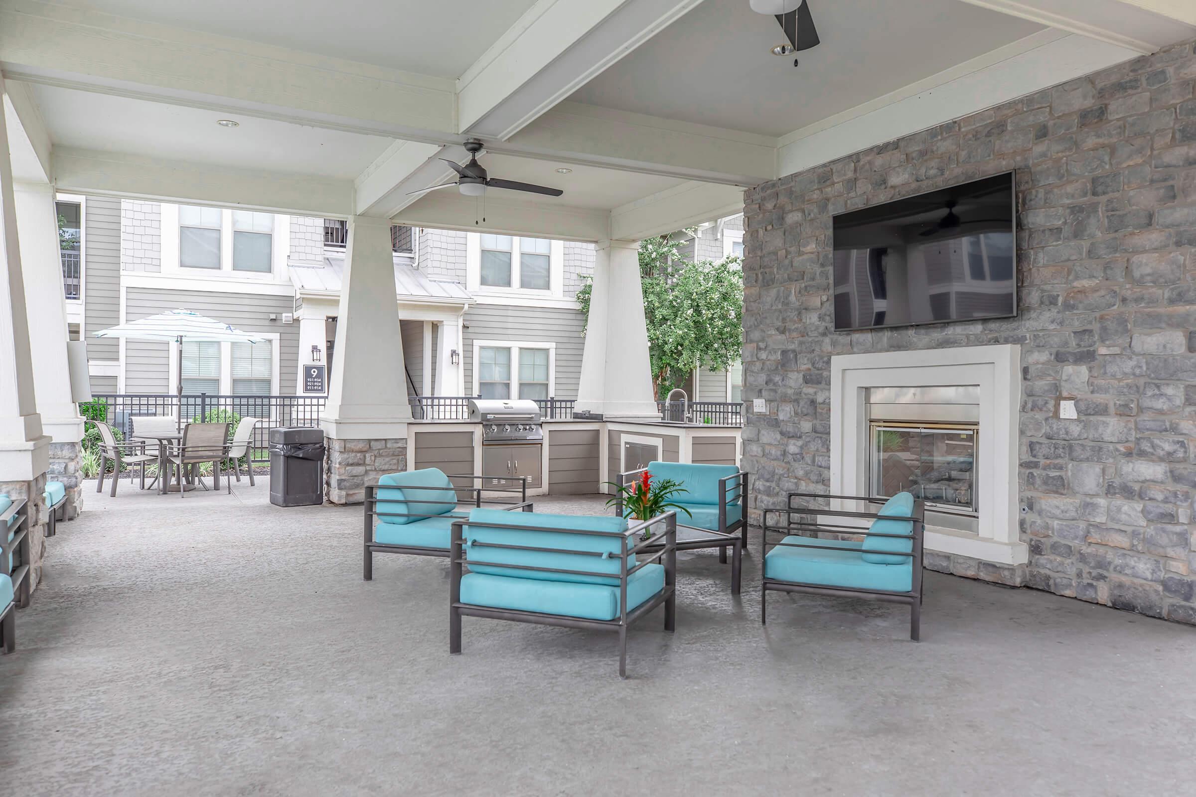 Outdoor seating area featuring modern blue chairs and a table, surrounded by stone walls. A wall-mounted TV is visible, along with a fireplace. The space is well-lit and overlooks an outdoor area with trees and additional seating in the background.