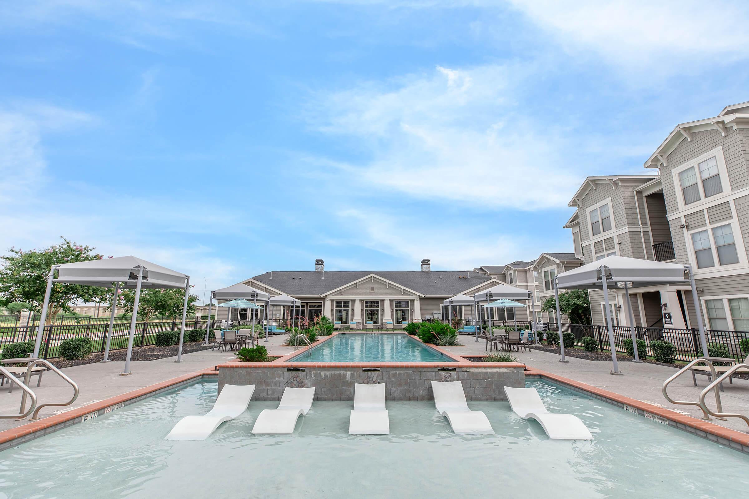 A modern apartment complex featuring a spacious outdoor pool with lounge chairs and shaded cabanas. The pool area is surrounded by lush greenery and stylish architectural details of the buildings in the background, set against a clear blue sky.