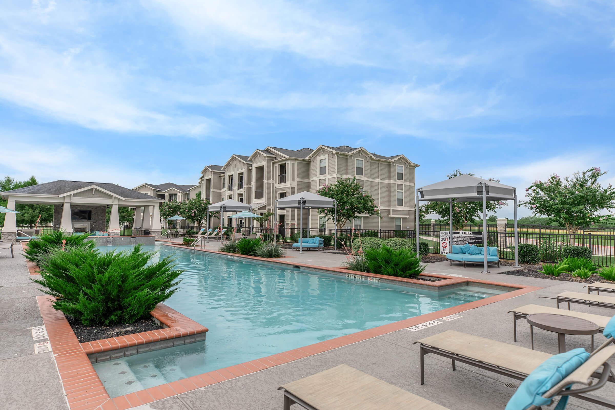 A tranquil outdoor pool area surrounded by lounge chairs, umbrellas, and lush greenery. In the background, a modern apartment building is visible against a clear blue sky. The space is inviting and designed for relaxation.
