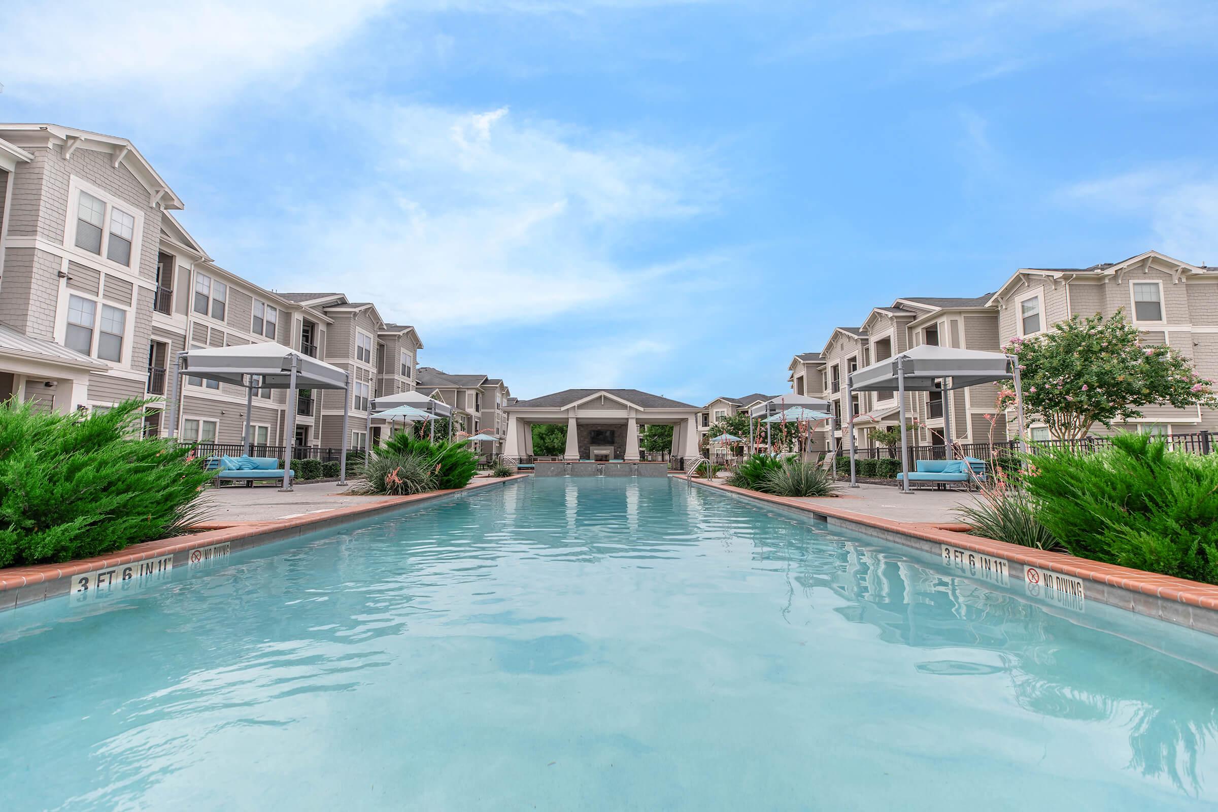 A tranquil swimming pool surrounded by lush greenery and modern apartment buildings. The water is clear, reflecting a bright blue sky. Umbrellas and seating areas are visible along the poolside, creating a relaxing atmosphere for residents and guests.