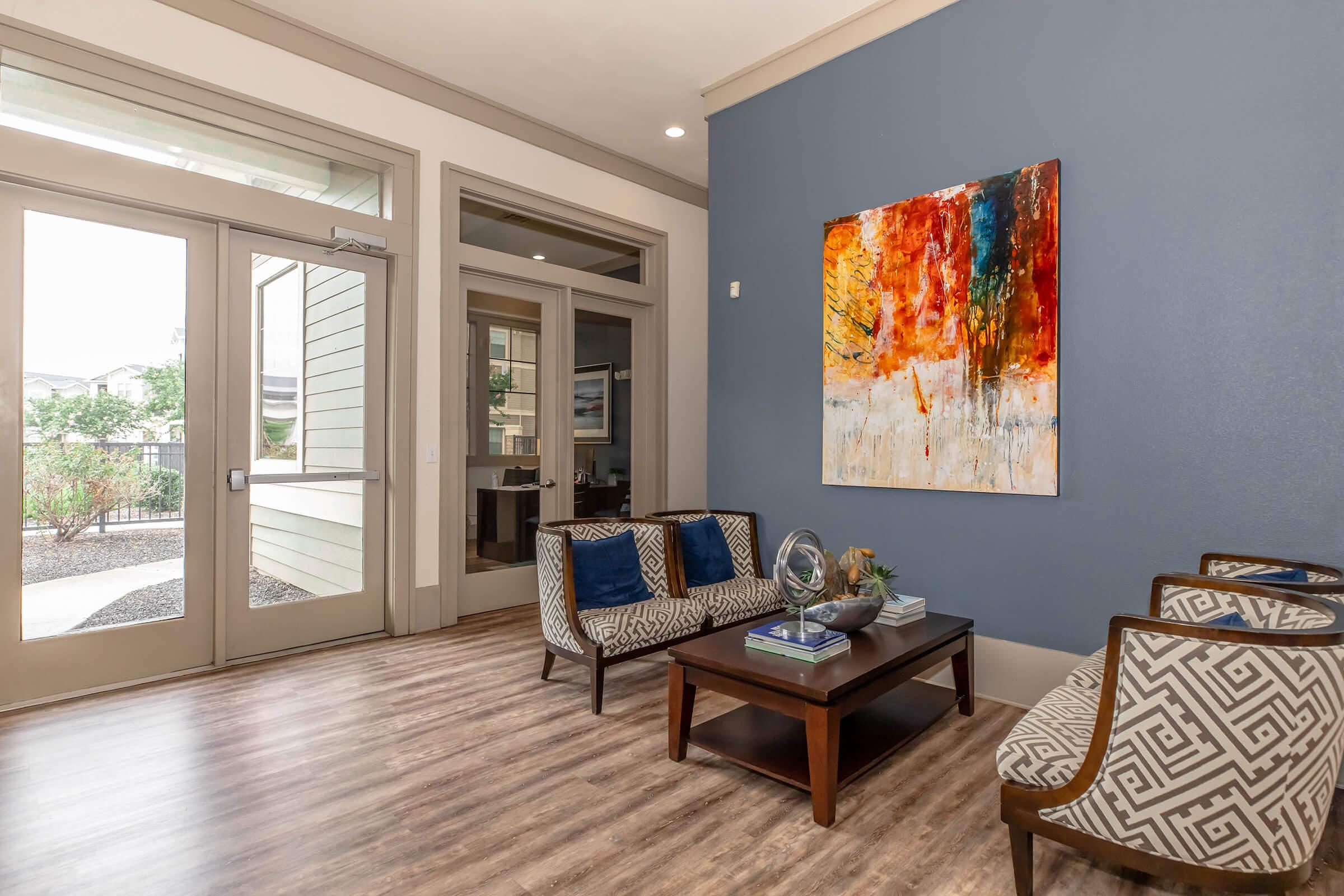 A modern interior space featuring a seating area with patterned chairs and a wooden coffee table. The room has large glass doors leading outside and a colorful abstract painting on the wall. Natural light illuminates the area, enhancing the warm wood flooring and contemporary decor.