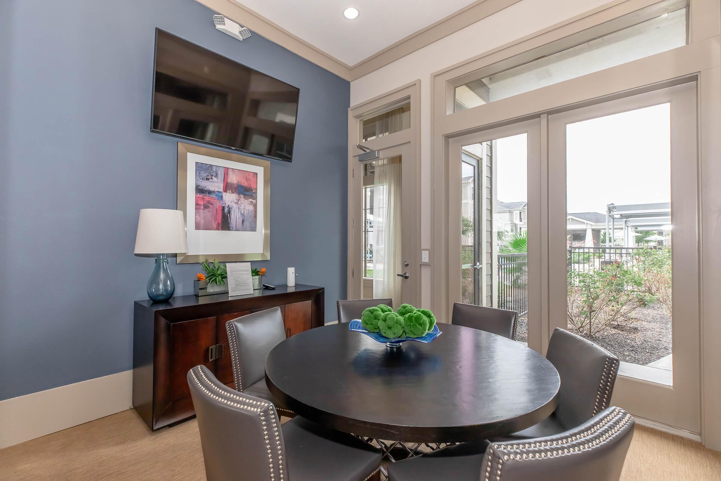 A modern room featuring a round black table surrounded by gray chairs, a stylish lamp, and a cabinet. A colorful abstract artwork hangs on the wall beneath a mounted television. Large windows offer a view of outdoor greenery, allowing natural light to illuminate the space.