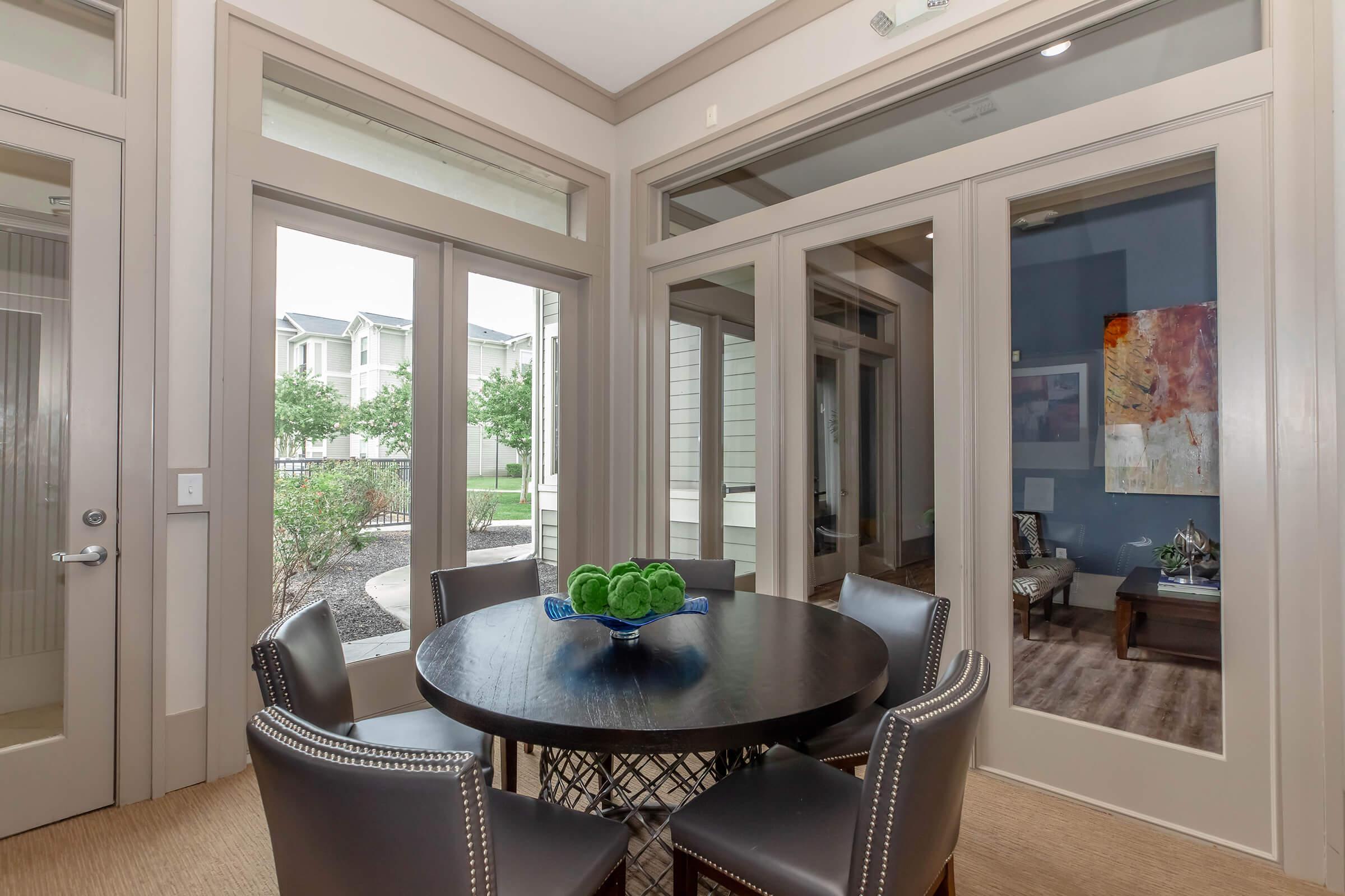 A modern dining area with a round table surrounded by high-backed chairs. The room features large windows allowing natural light, and a decorative bowl with green items on the table. A view of greenery through the windows can be seen, along with a stylish side table and artwork on the wall.
