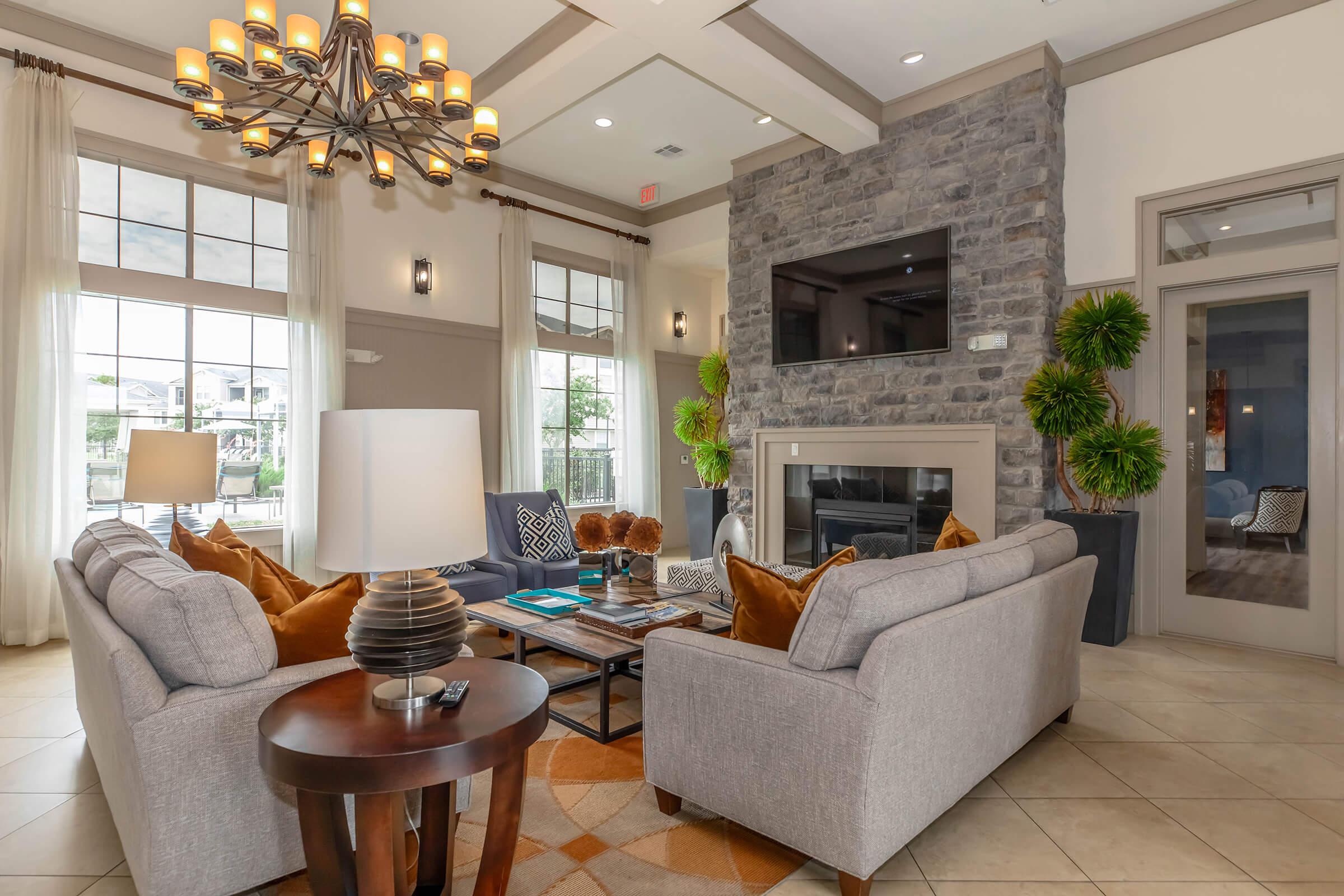 A cozy living room with gray sofas adorned with orange and brown cushions, a stylish round table, and a modern chandelier. A stone fireplace is the focal point, complemented by large windows letting in natural light and potted plants adding greenery to the space. A television is mounted on the wall.