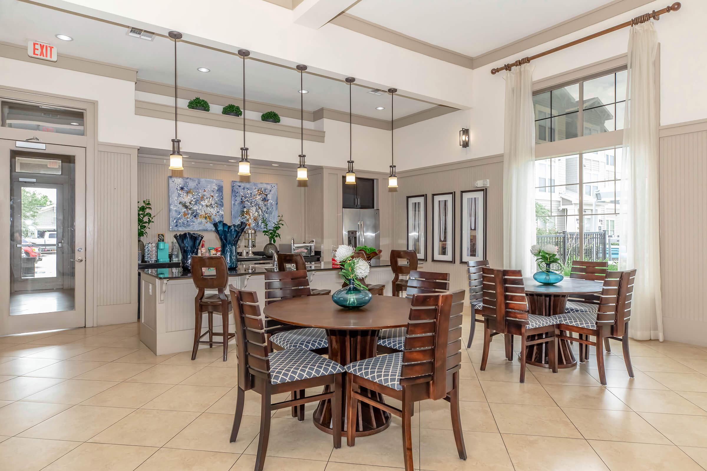 Modern dining area featuring dark wood tables and chairs arranged around circular tables. The space includes large windows allowing natural light, pendant lighting, and decorative plants. A kitchen area is visible in the background with cabinetry and a blue artwork display.