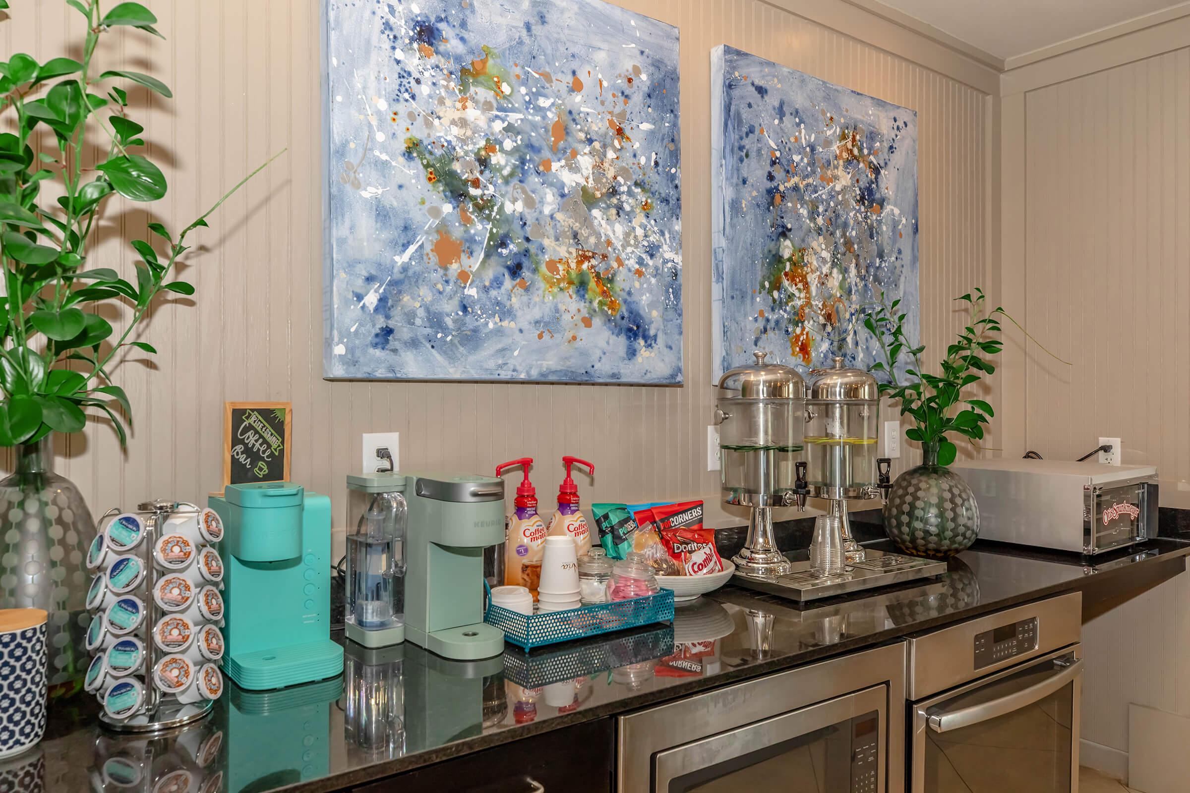 A stylish kitchen bar area featuring two abstract paintings in blue and gold tones. On the counter, there are a coffee maker, water dispenser, flavored syrups, and a variety of coffee pods. Decorative plants and containers enhance the aesthetic, complemented by a sleek stainless steel microwave and fridge.