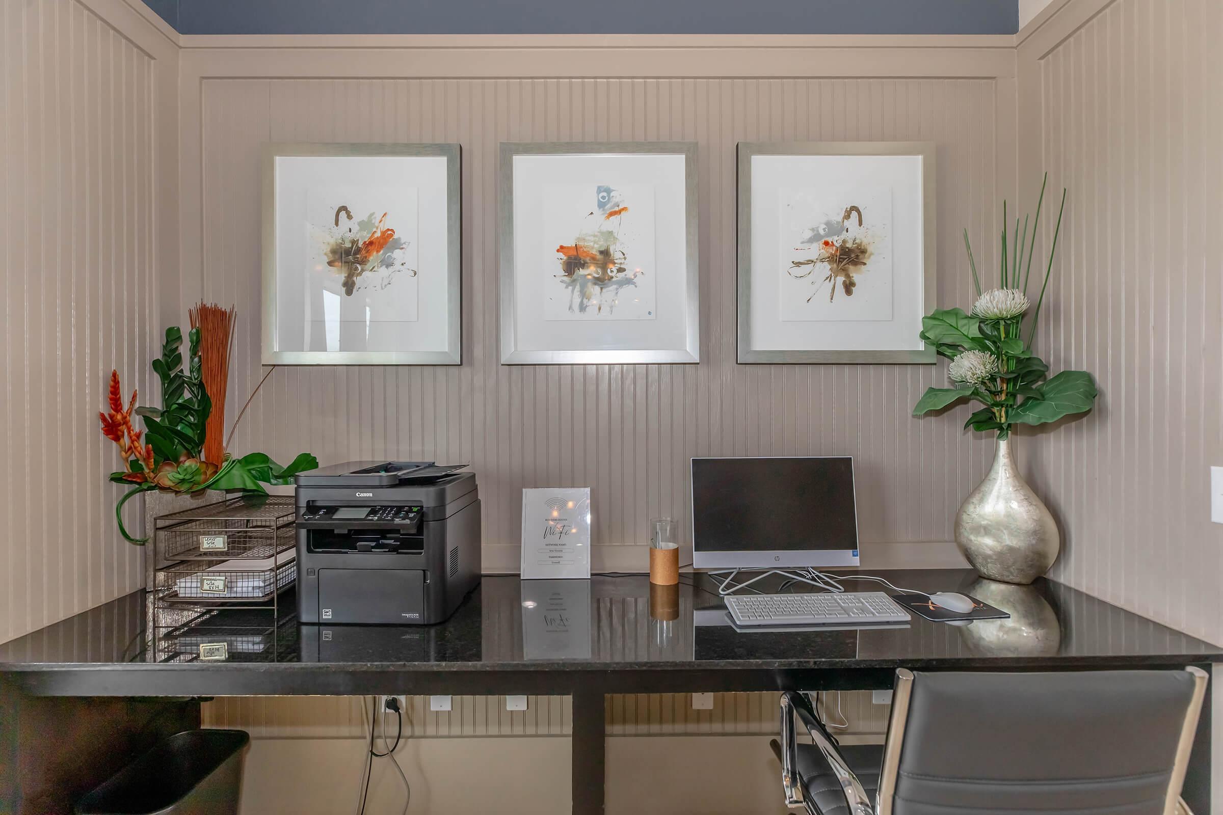 A modern home office setup featuring a sleek black desk with a computer monitor, keyboard, and printer. The wall is adorned with three framed abstract art pieces. A decorative vase with greenery and a small potted plant add a touch of nature, while organized storage boxes sit neatly on the desk.