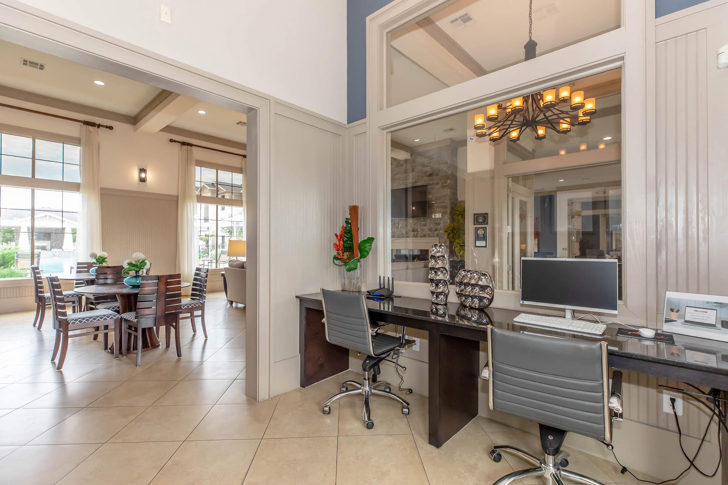 A modern office space with a dark desk and ergonomic chairs. A computer is visible on the desk, alongside decorative items. In the background, a bright dining area features a wooden dining table and chairs, with large windows letting in natural light, creating an inviting atmosphere.