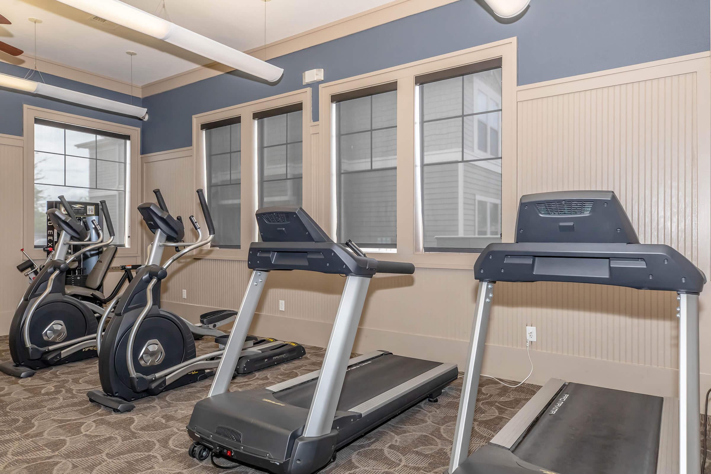 A well-lit gym area featuring multiple exercise machines including treadmills and an elliptical trainer. The room has large windows with blinds, beige and blue walls, and a plush carpet, creating a modern and inviting atmosphere for workouts.