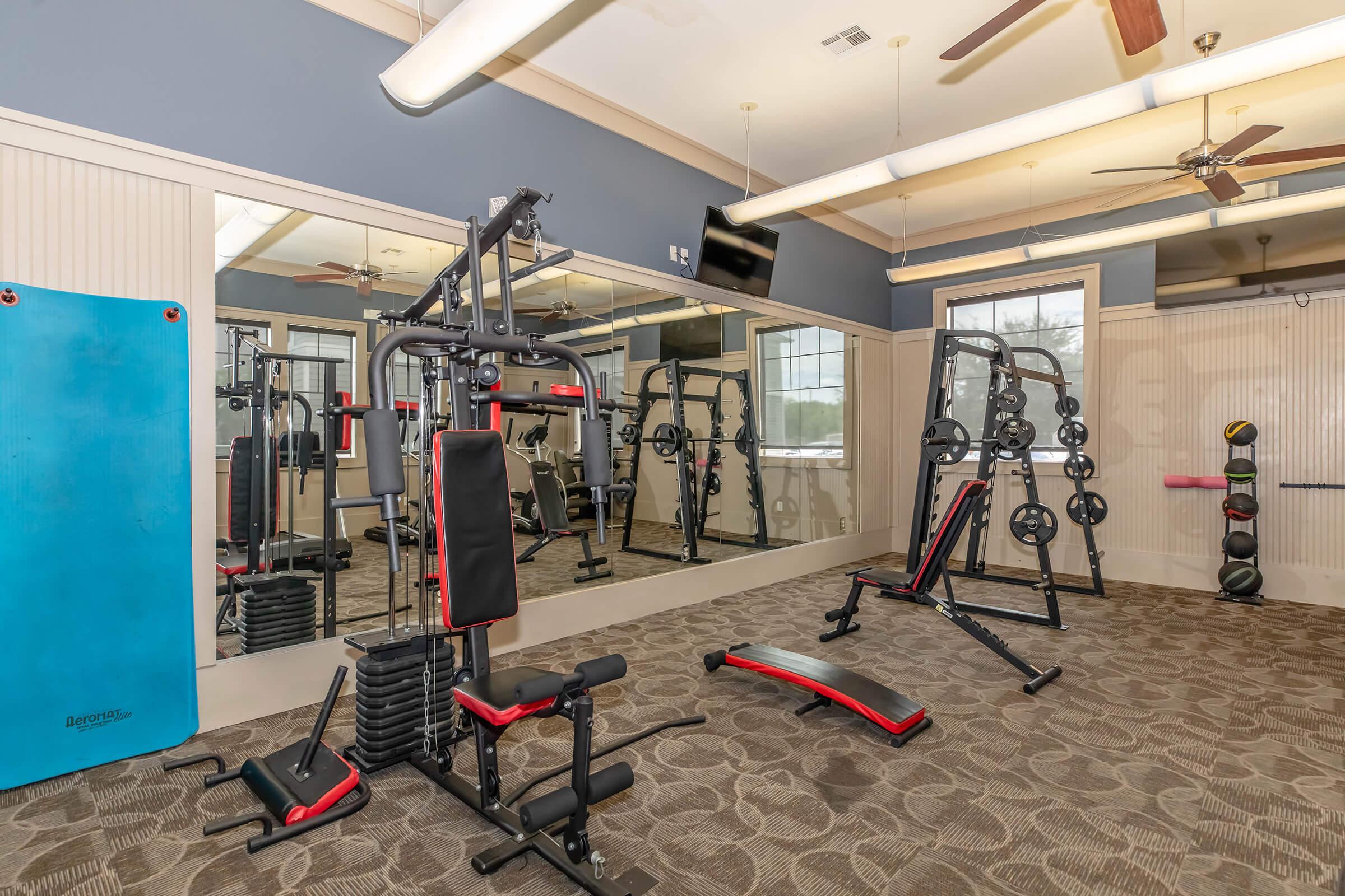 A spacious gym area featuring exercise equipment including a multi-station weight machine, a bench, free weights, and a set of resistance bands on the wall. Large mirrors line one wall, reflecting the equipment and a well-lit, airy environment.