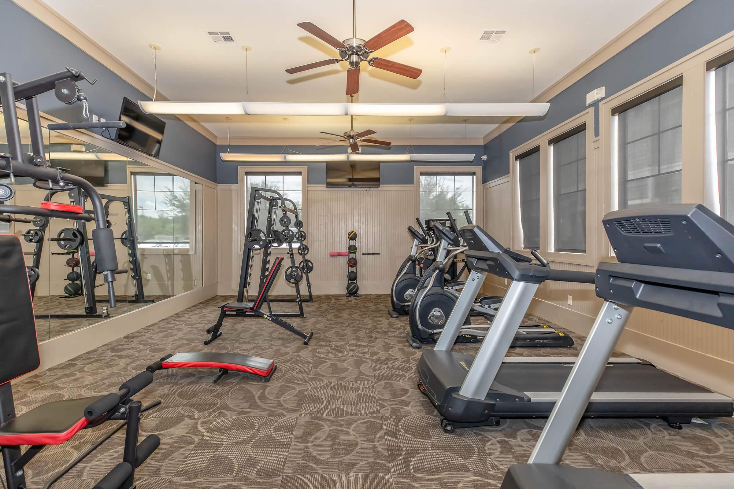 A modern fitness room featuring various gym equipment including treadmills, weight machines, and benches. The space is well-lit with ceiling fans and large windows, giving a bright and open feel. A mirror line one wall, enhancing the workout atmosphere.