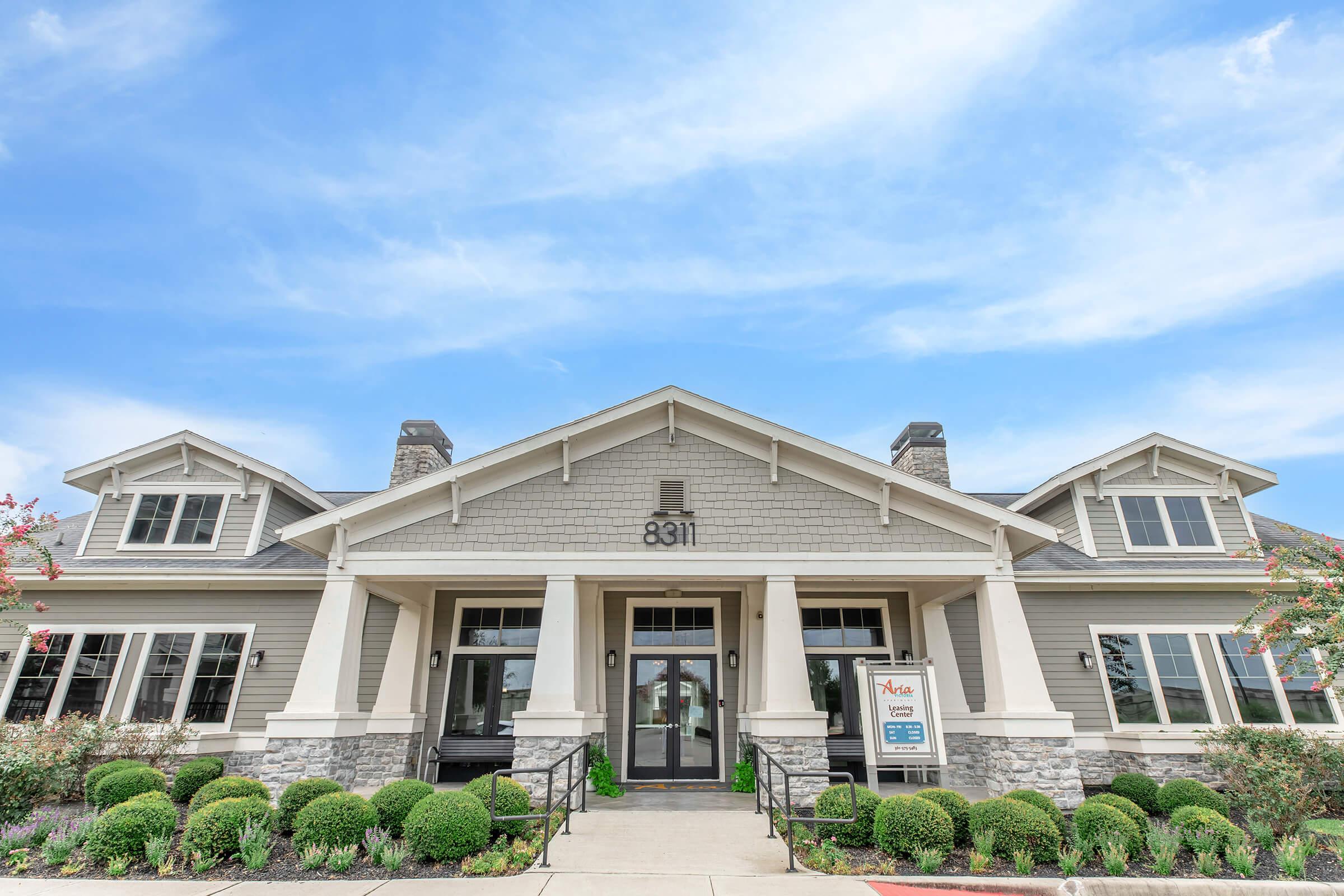 A modern, two-story building with a gray and white exterior, featuring a prominent entrance with large glass doors and decorative stone accents. The facade includes a street number "8311" and is surrounded by neatly trimmed bushes and landscaping under a bright blue sky.