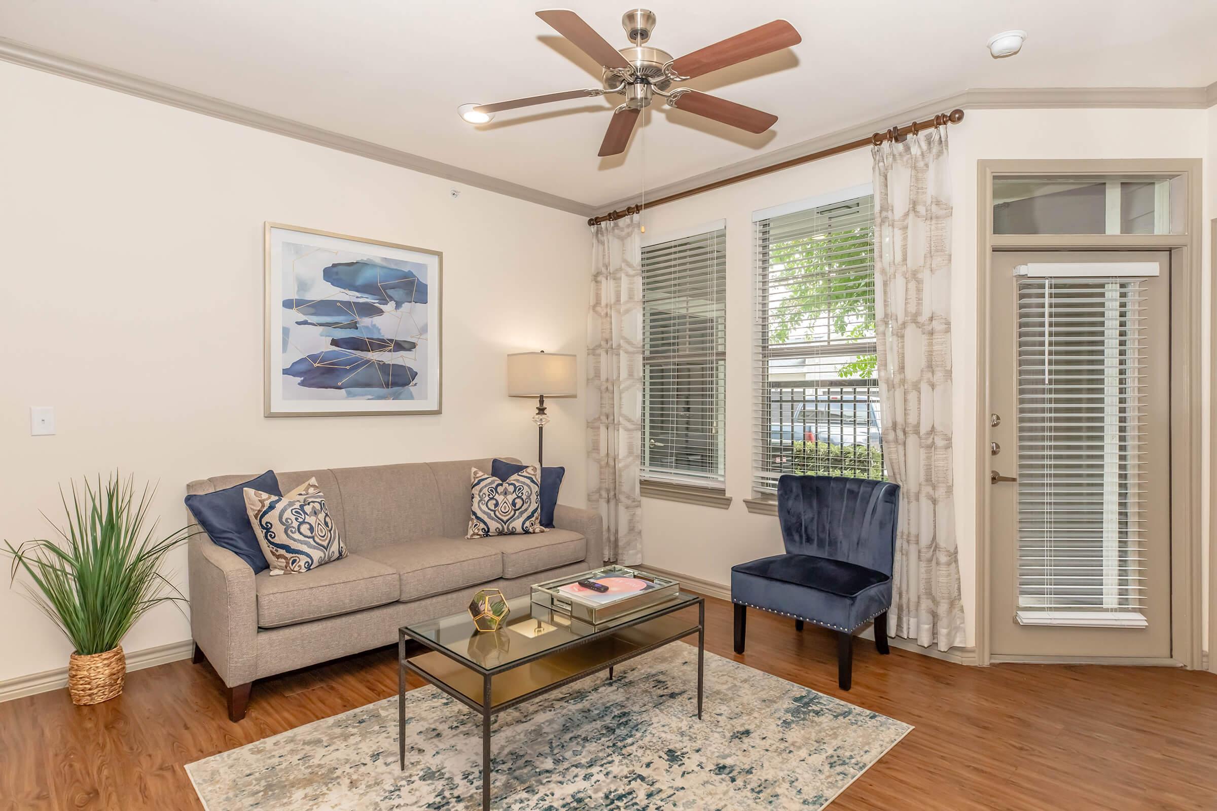 A cozy living room featuring a neutral-colored sofa with decorative pillows, a glass coffee table, and a navy blue accent chair. Natural light streams in through large windows with light curtains. A piece of artwork depicting boats adorns the wall, and a small plant adds a touch of greenery.