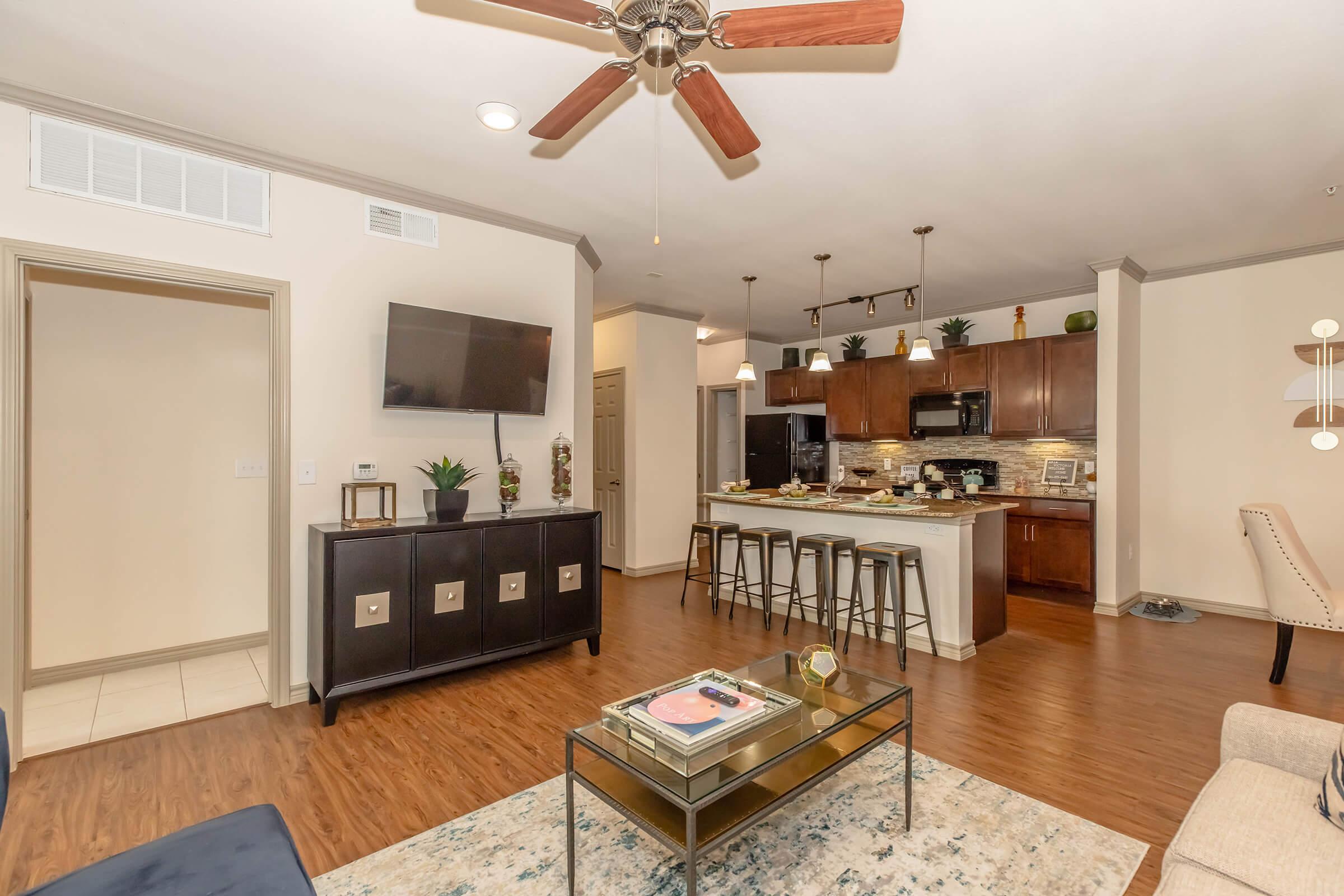 A modern living room with a ceiling fan, a flat-screen TV mounted on the wall, and stylish furniture. The open kitchen features dark wood cabinets and bar stools at an island. Natural light illuminates the space, which includes a decorative rug and a coffee table. An adjacent room with a closed door is visible.