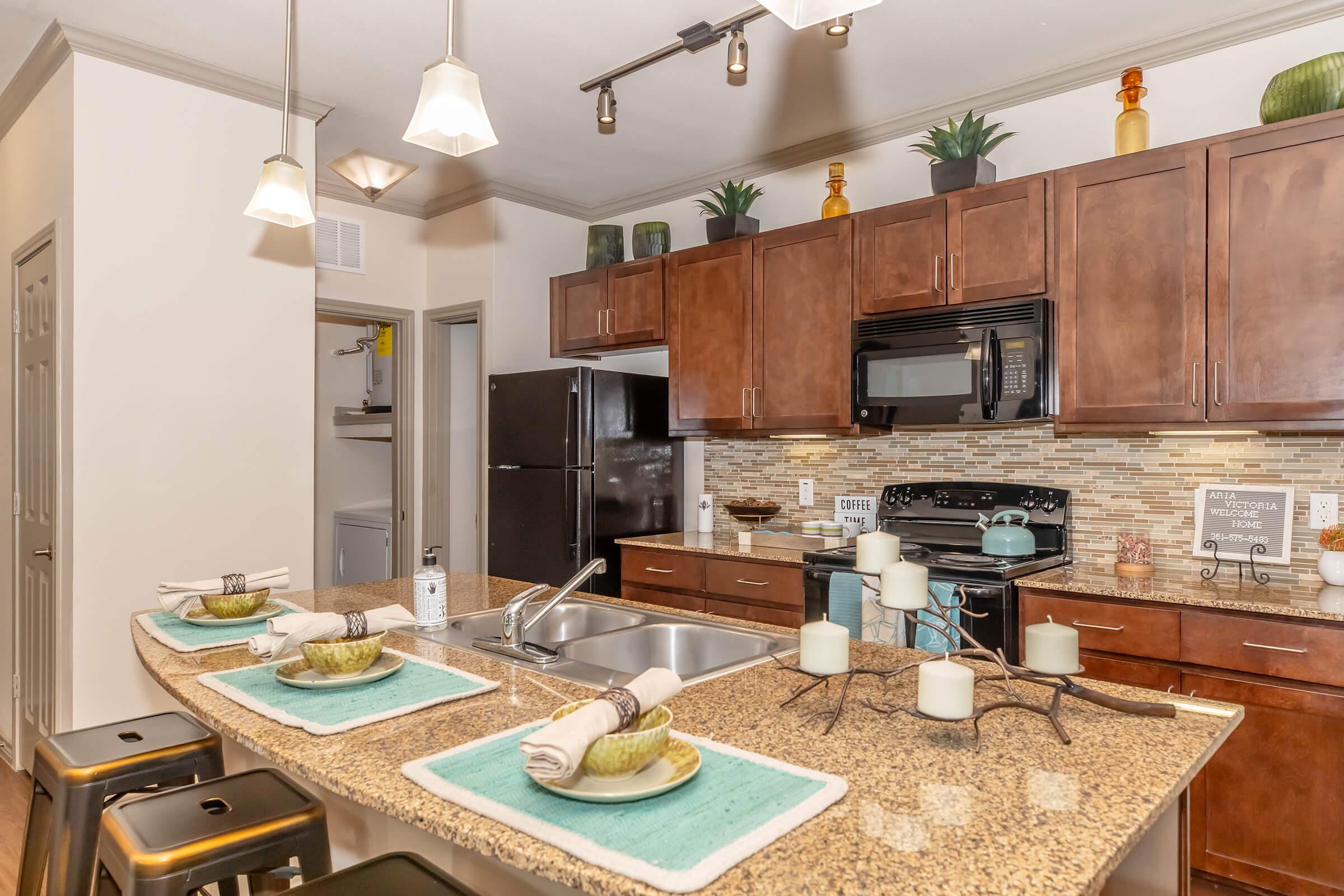 Modern kitchen featuring dark wood cabinets, granite countertops, and a tile backsplash. Equipped with stainless steel appliances, including a refrigerator and microwave. There are bar stools at the island with decorative plates and candles. Light fixtures hang from the ceiling, and green plants are arranged decoratively on the shelves.