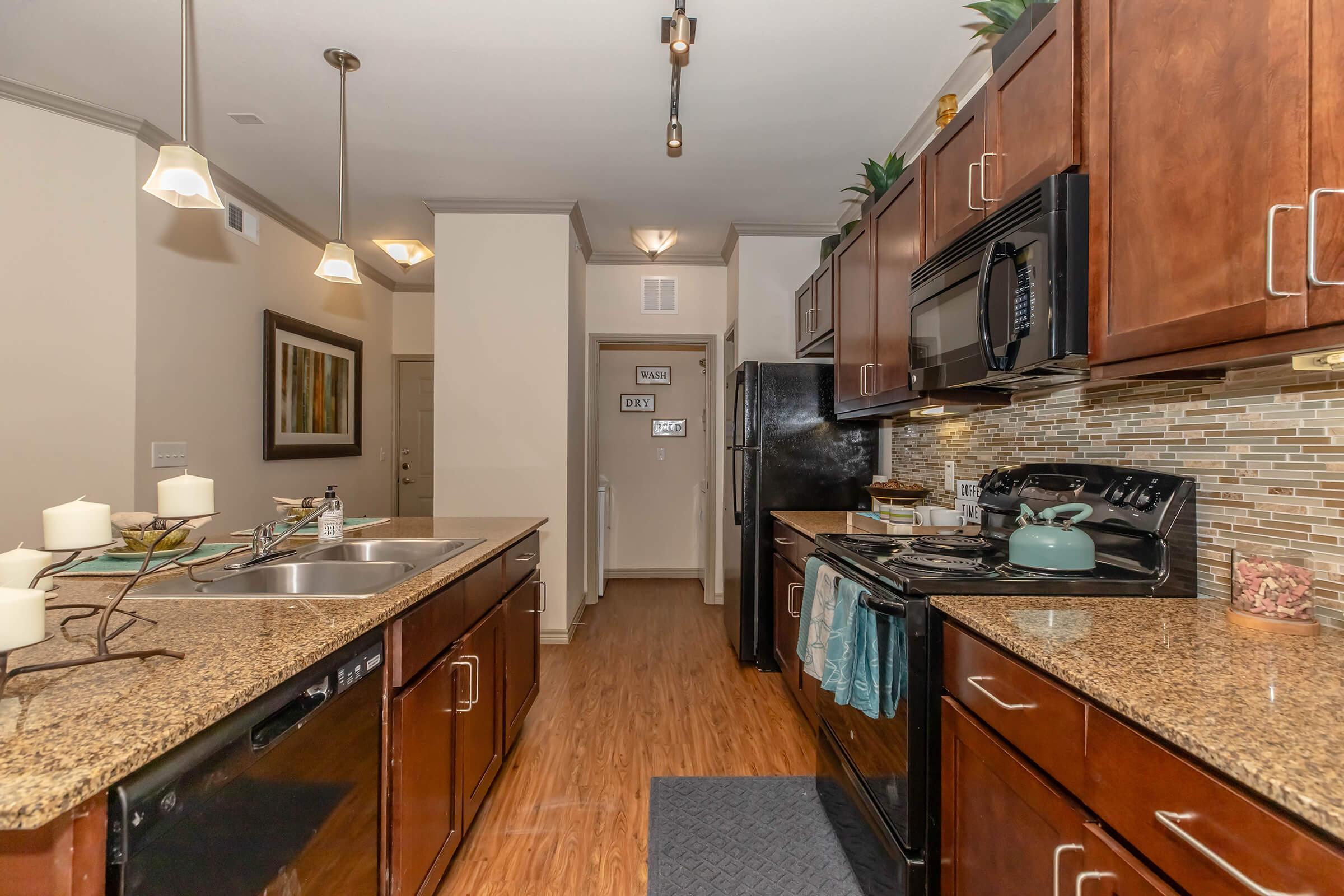 Modern kitchen featuring granite countertops, dark wooden cabinetry, and stainless steel appliances. The space includes a double sink, a gas stove, and decorative accents like potted plants and candles. Light fixtures hang above, and a doorway leads to another room in the background.