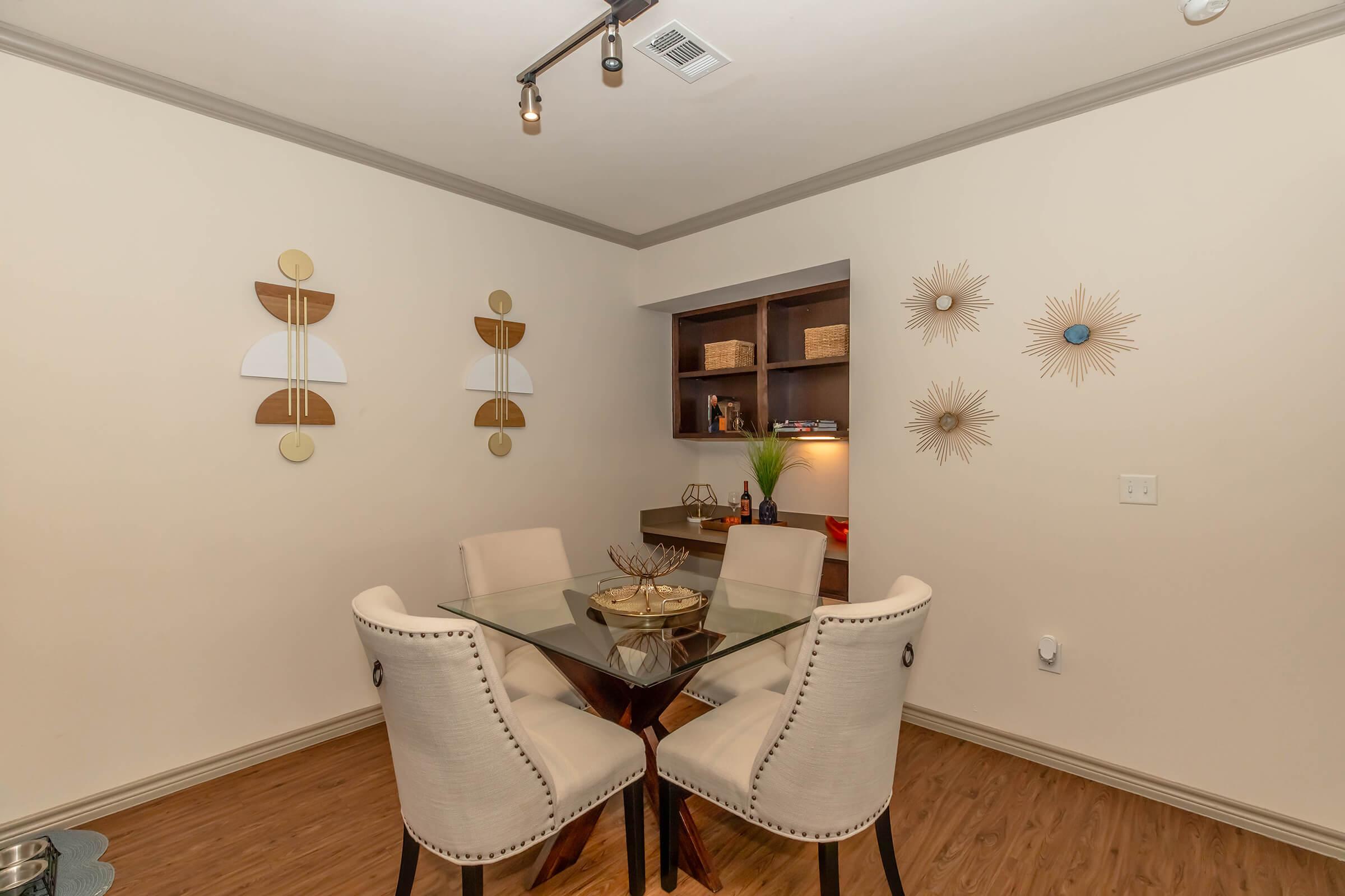 A stylish dining area featuring a glass dining table surrounded by four upholstered chairs. The walls are adorned with decorative wall art, including two light wood and white pieces and three starburst-shaped decorations. A small bookshelf displays decorative items, adding a modern touch to the space.