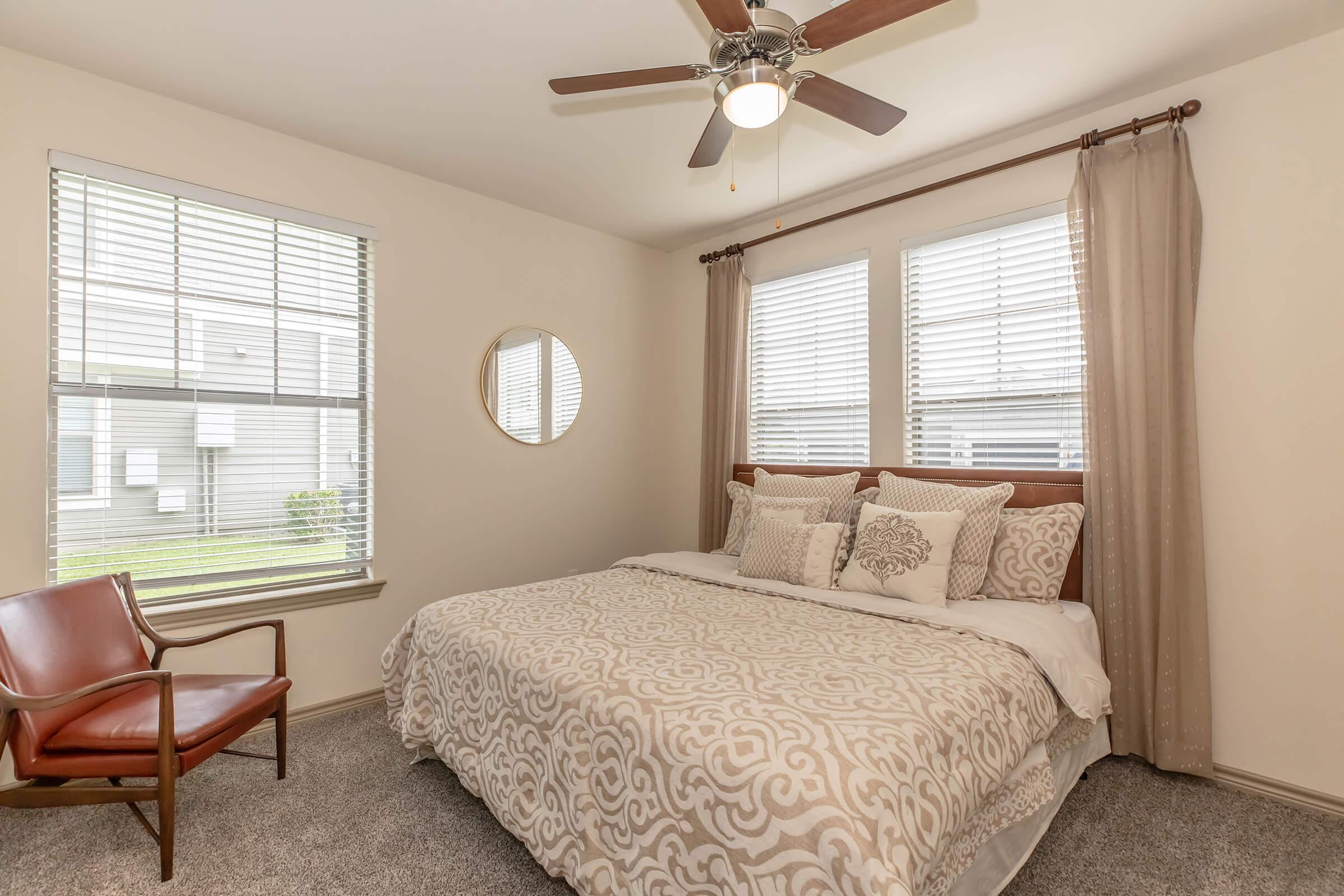 A cozy bedroom featuring a large bed with decorative pillows, a round mirror on the wall, and two windows with blinds allowing natural light. An armchair is situated beside the bed, and the room has light-colored walls and gray carpet flooring, creating a warm and inviting atmosphere.