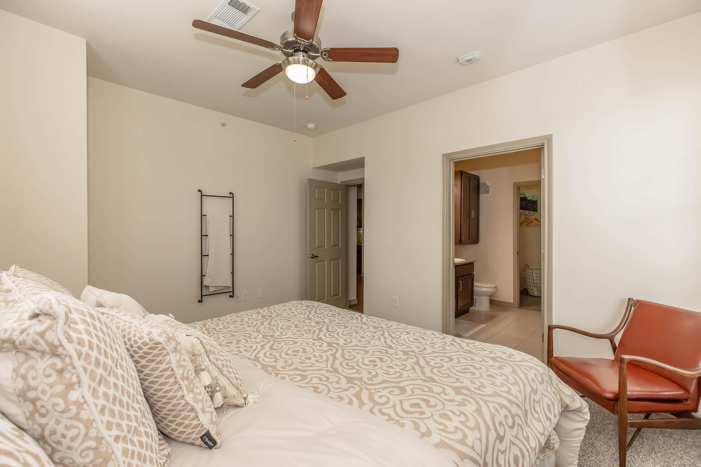 A cozy bedroom featuring a large bed with decorative pillows, a ceiling fan, and a modern chair. The room has warm beige walls, a full-length mirror on the wall, and an open doorway leading to a bathroom. Soft lighting creates a welcoming atmosphere.