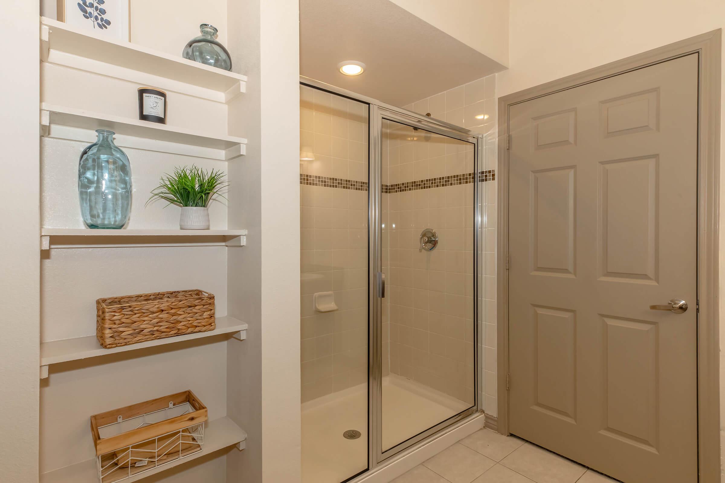 Bathroom interior featuring a glass shower stall, beige walls, and a door. A corner shelf displays decorative items, including vases and a plant. The tile floor complements the neutral color scheme.