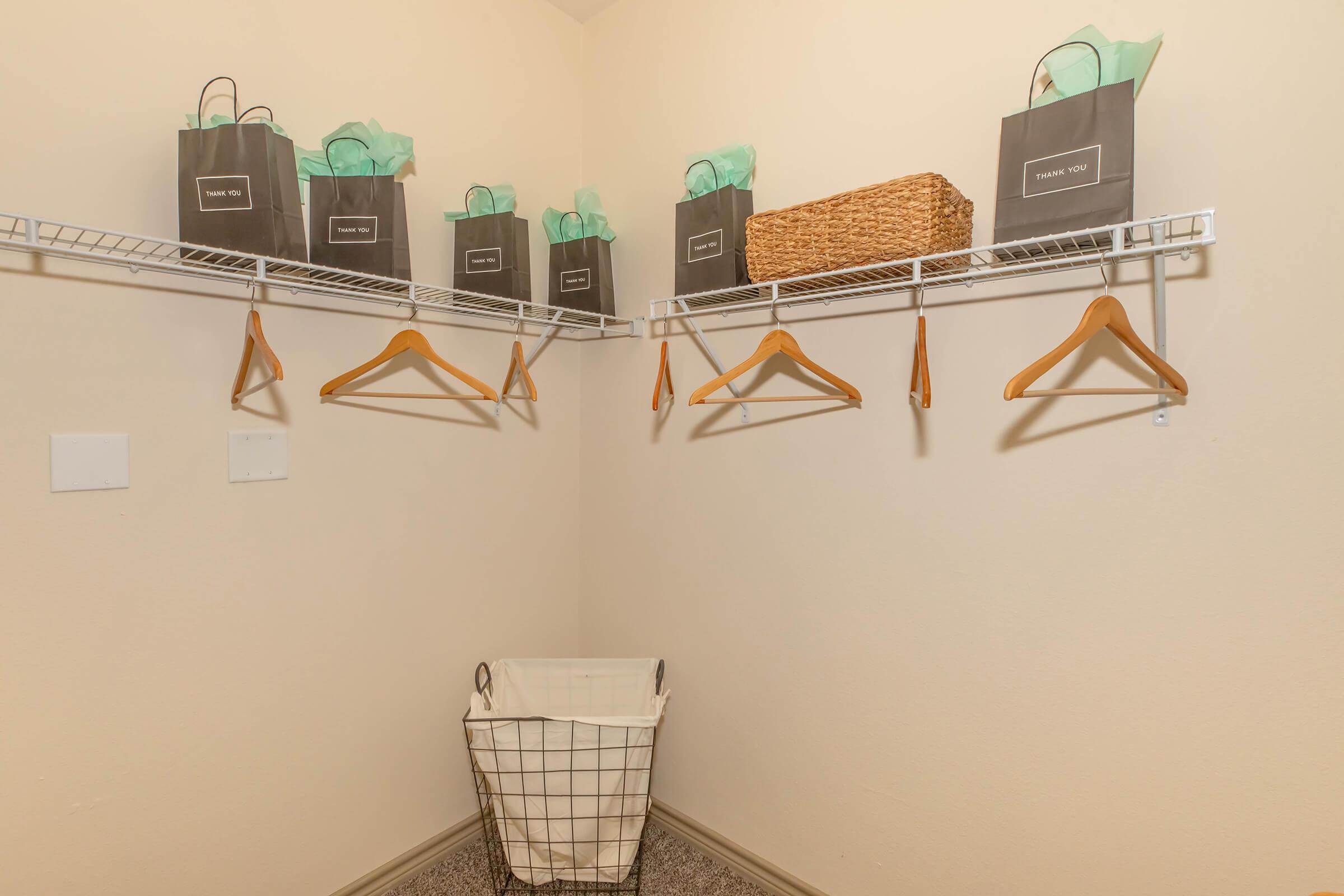 A neat closet with a light-colored wall featuring a wire shelf. On the shelf, there are several black gift bags with turquoise tissue paper, a woven basket, and a few wooden hangers hanging from the rod. A wire laundry basket sits on the floor, adding to the organized appearance.