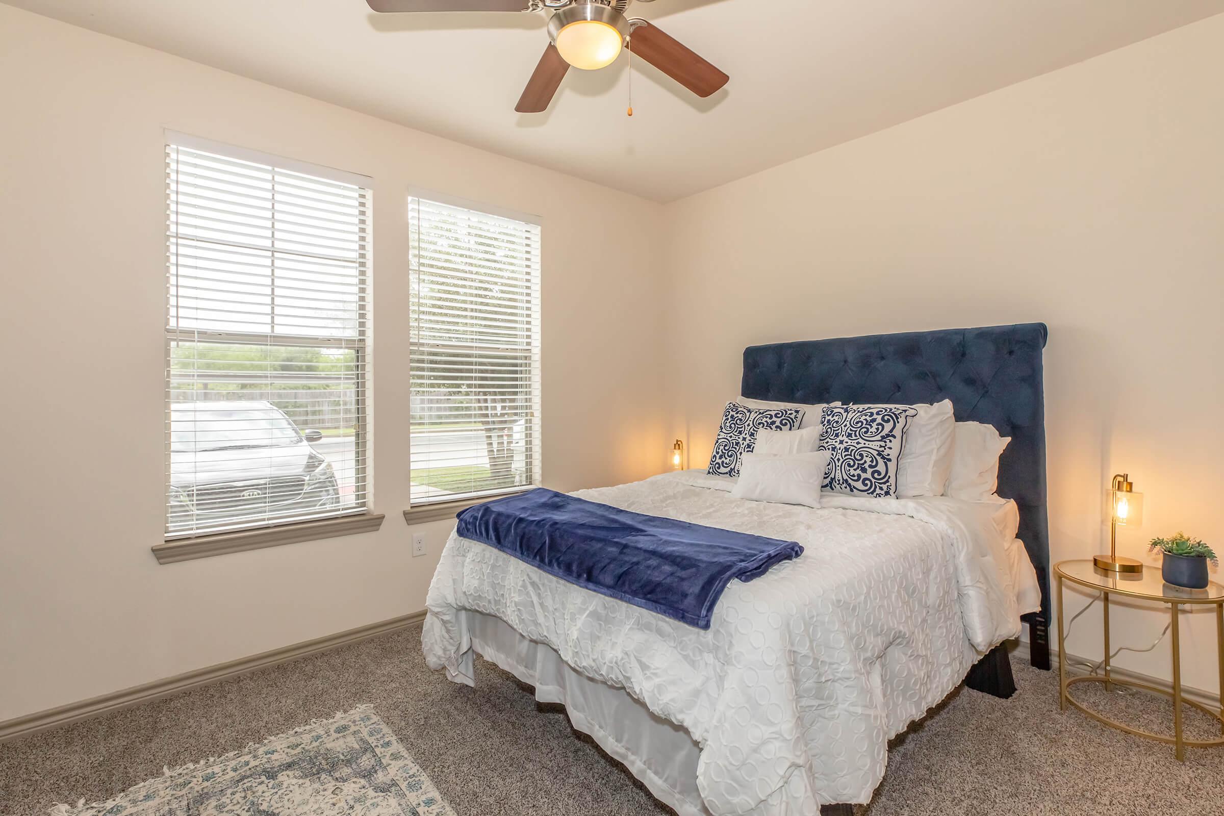 A cozy bedroom featuring a blue upholstered headboard bed with white bedding, decorative pillows, and a blue throw blanket. There are two windows allowing natural light, and a small side table with a plant and lamp beside the bed. The room has a soft carpet and neutral-colored walls.