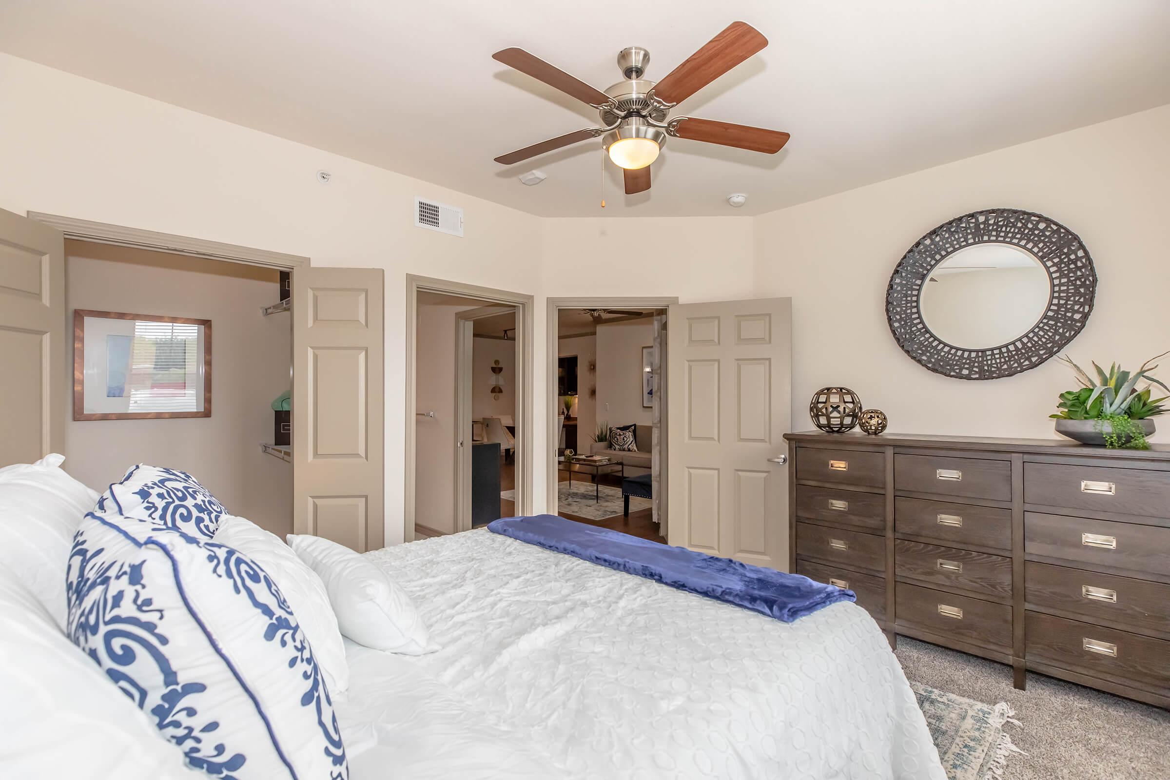 Cozy bedroom featuring a large bed with decorative pillows, a ceiling fan, and a stylish mirror above a dresser. Two doorways lead to other rooms, and a small plant adds a touch of greenery. Natural light filters through the window, enhancing the warm, inviting atmosphere.