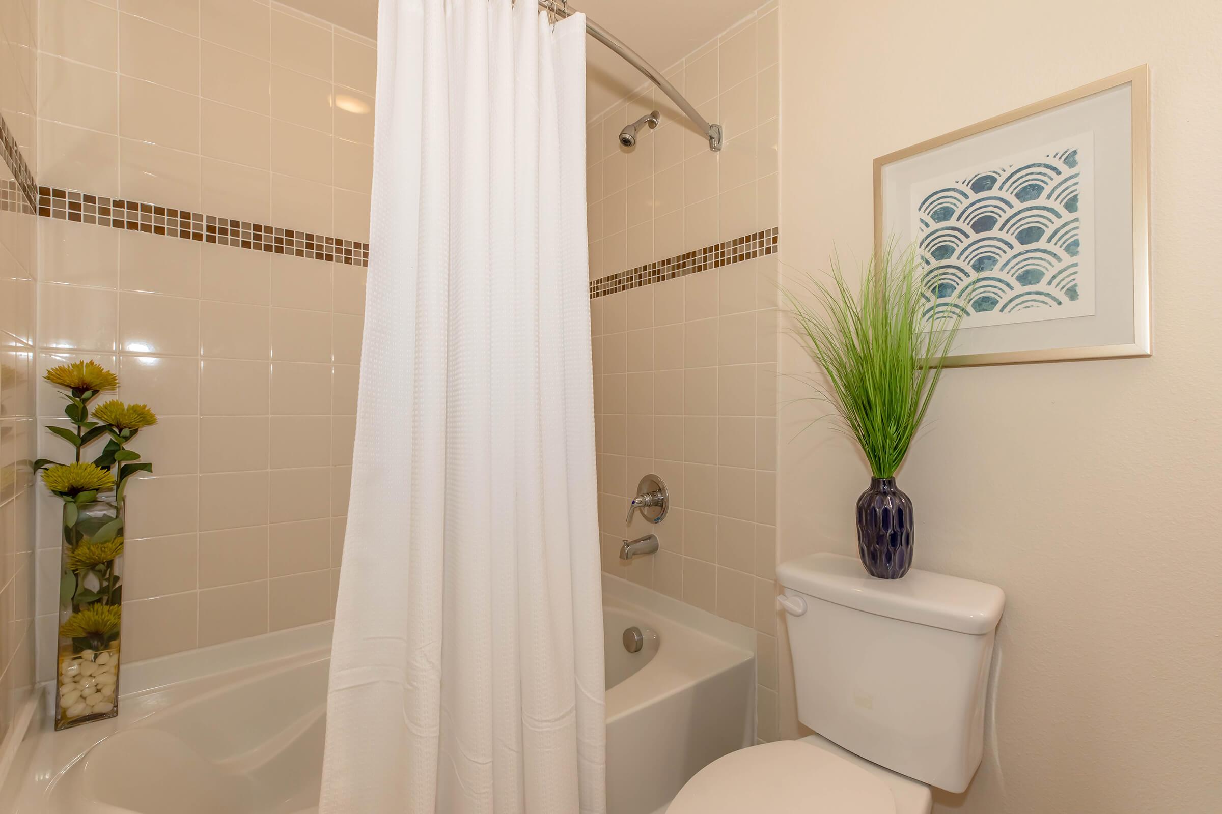 A clean bathroom featuring a bathtub with a shower curtain, a toilet, and a decorative vase with faux plants. The walls are tiled in light colors, with a framed artwork hanging nearby. The space is well-lit and organized, creating a welcoming atmosphere.