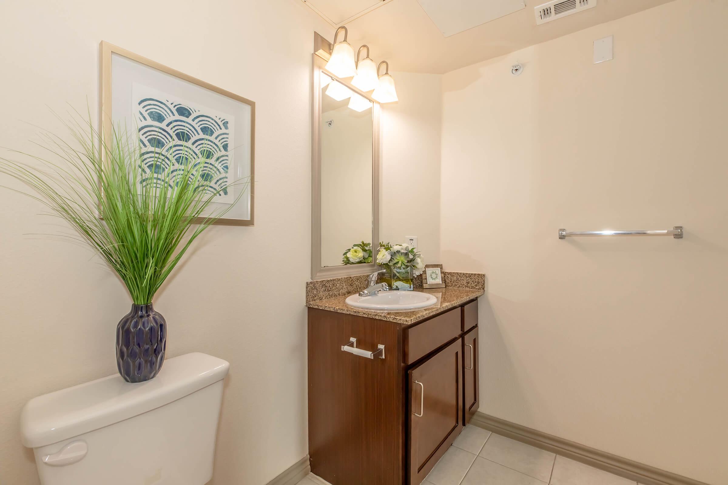 A well-lit bathroom featuring a small vanity with a sink and mirror, a decorative plant in a vase, a framed artwork on the wall, and a toilet. The color scheme includes neutral tones with a touch of greenery for decor.